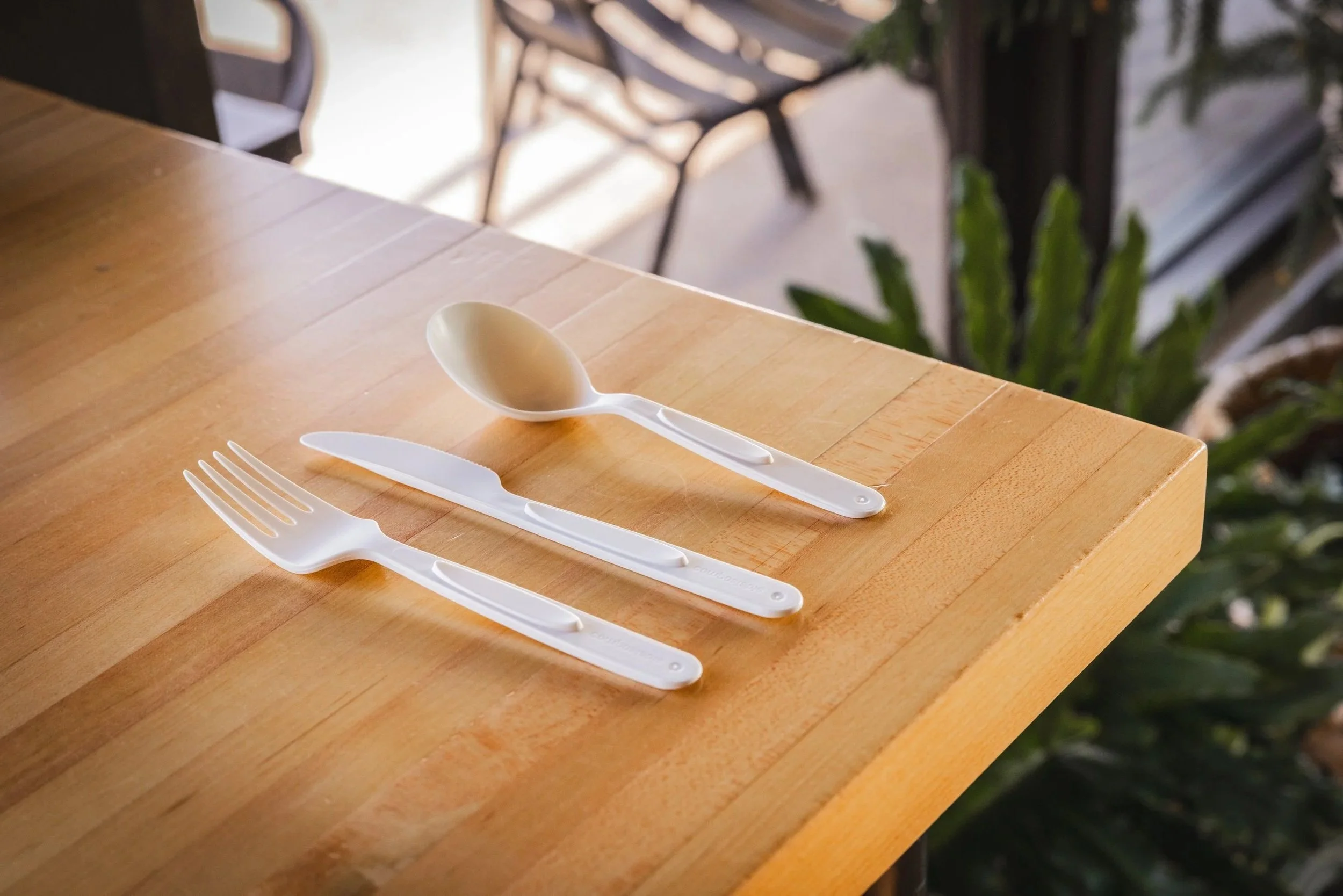 A wooden table with a set of disposable plastic utensils including a fork, knife, and spoon. The spoon is placed centrally, with the fork and knife beside it. The background is blurred, showing outdoor scenery with chairs, a table, and some green plants.
