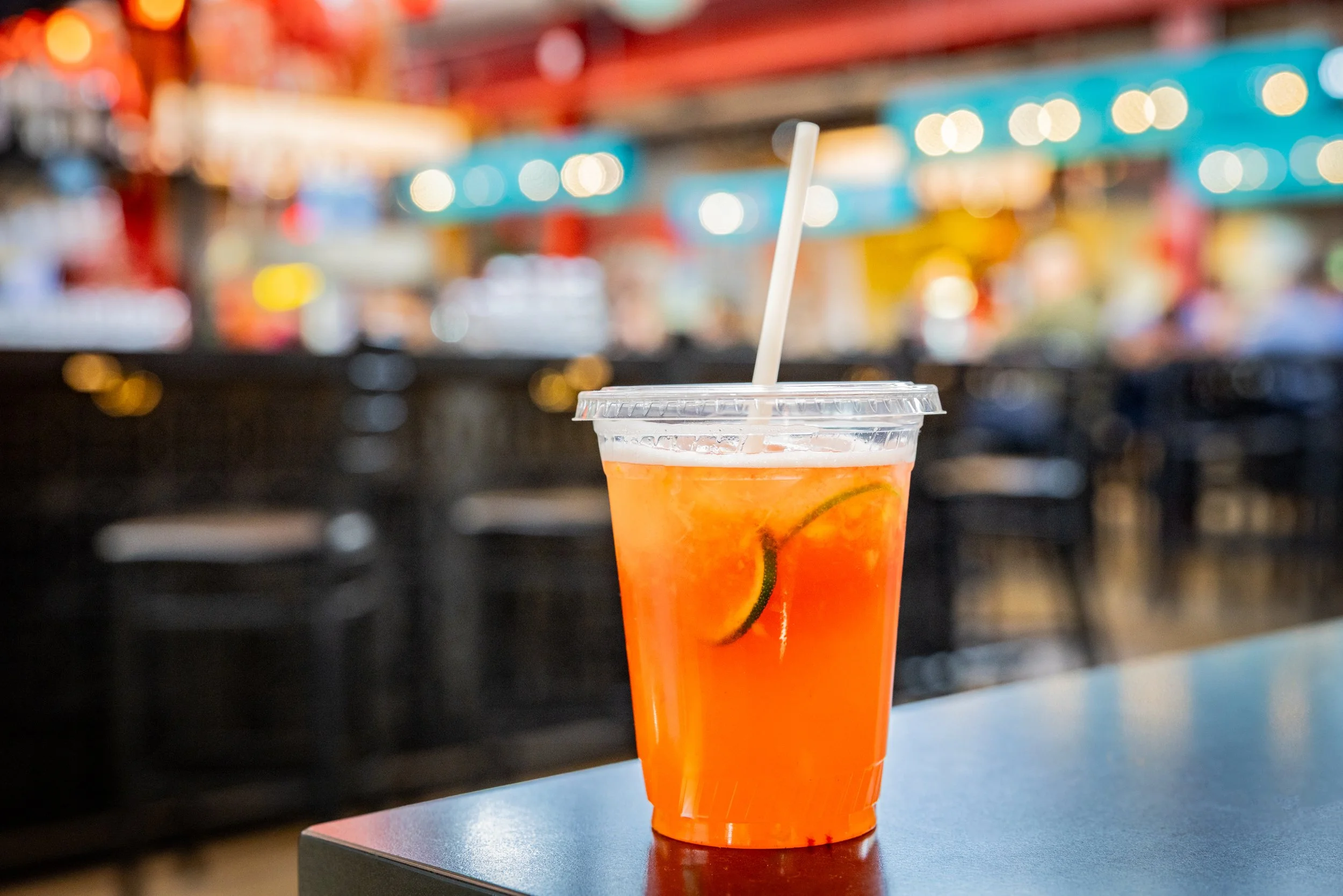 A plastic cup filled with an orange-colored beverage, garnished with slices of citrus fruit and a straw, on a dark table with a blurred, colorful background.
