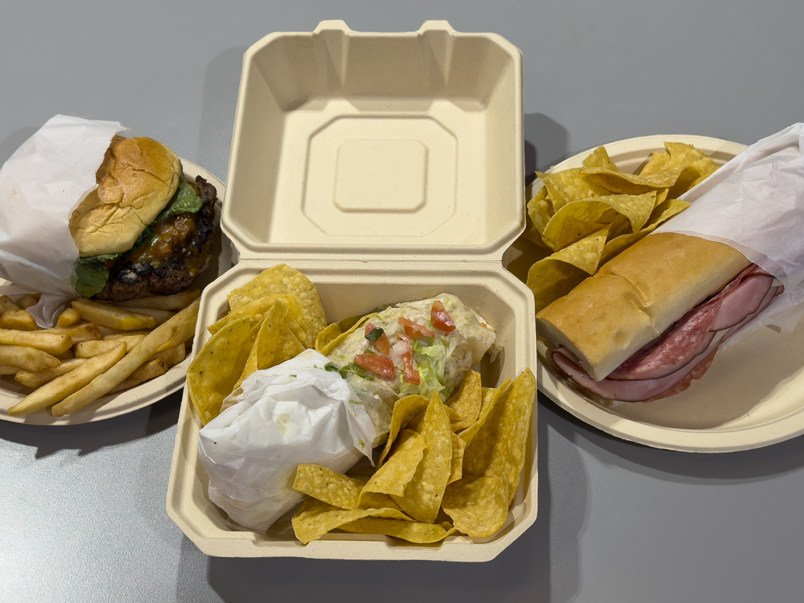 Two takeout containers and one plate with various fast food items including a burger with lettuce and sauce, a sub sandwich with cold cuts, and a portion of potato chips and tortilla chips, on a gray table.