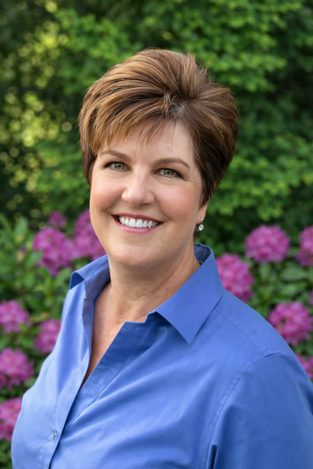 Portrait of a smiling woman with short brown hair, wearing a blue collared shirt, outdoors with greenery and purple flowers in the background.