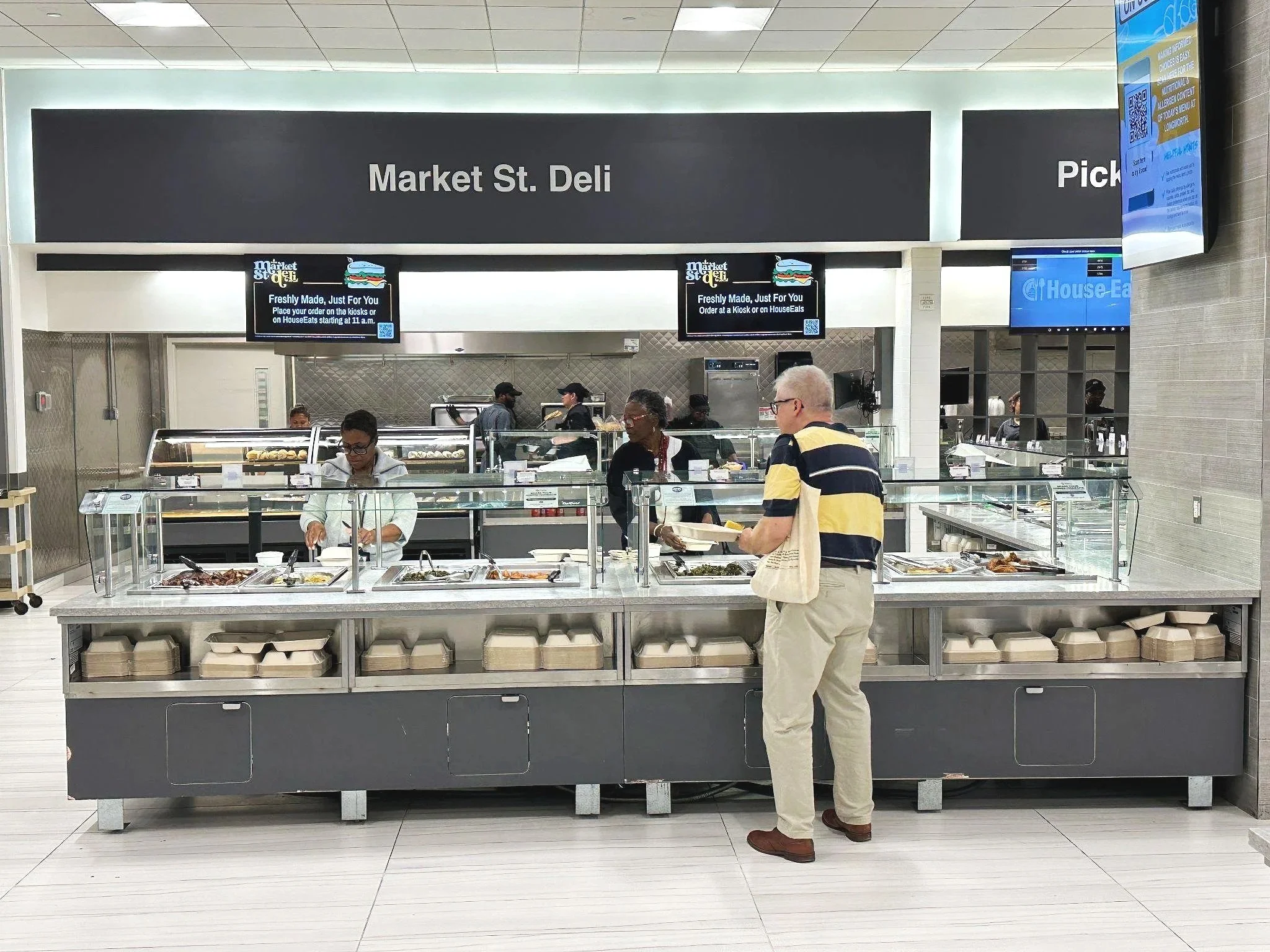 A man with glasses and a striped yellow and navy polo shirt holding a plate is standing at a food counter in a marketplace or food court. Behind the counter, staff members are serving food. The setting has a large black sign with white text reading 'Market St. Deli' and smaller digital screens displaying promotional messages. The decor is modern with light-colored walls and tiled floors.