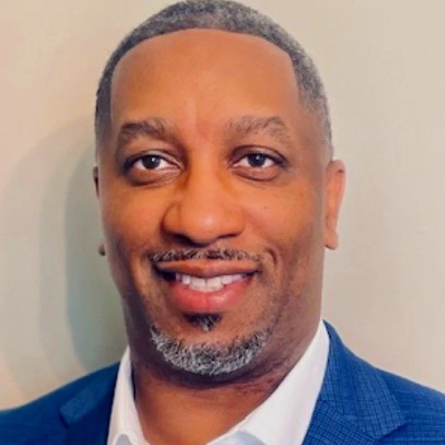 Close-up of a smiling man with short gray hair and a goatee, wearing a blue suit and white shirt, against a neutral background.