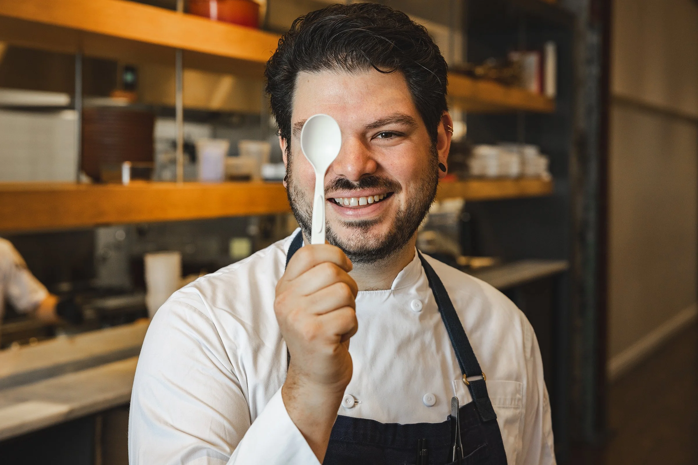A smiling male chef holding a white spoon in a kitchen.