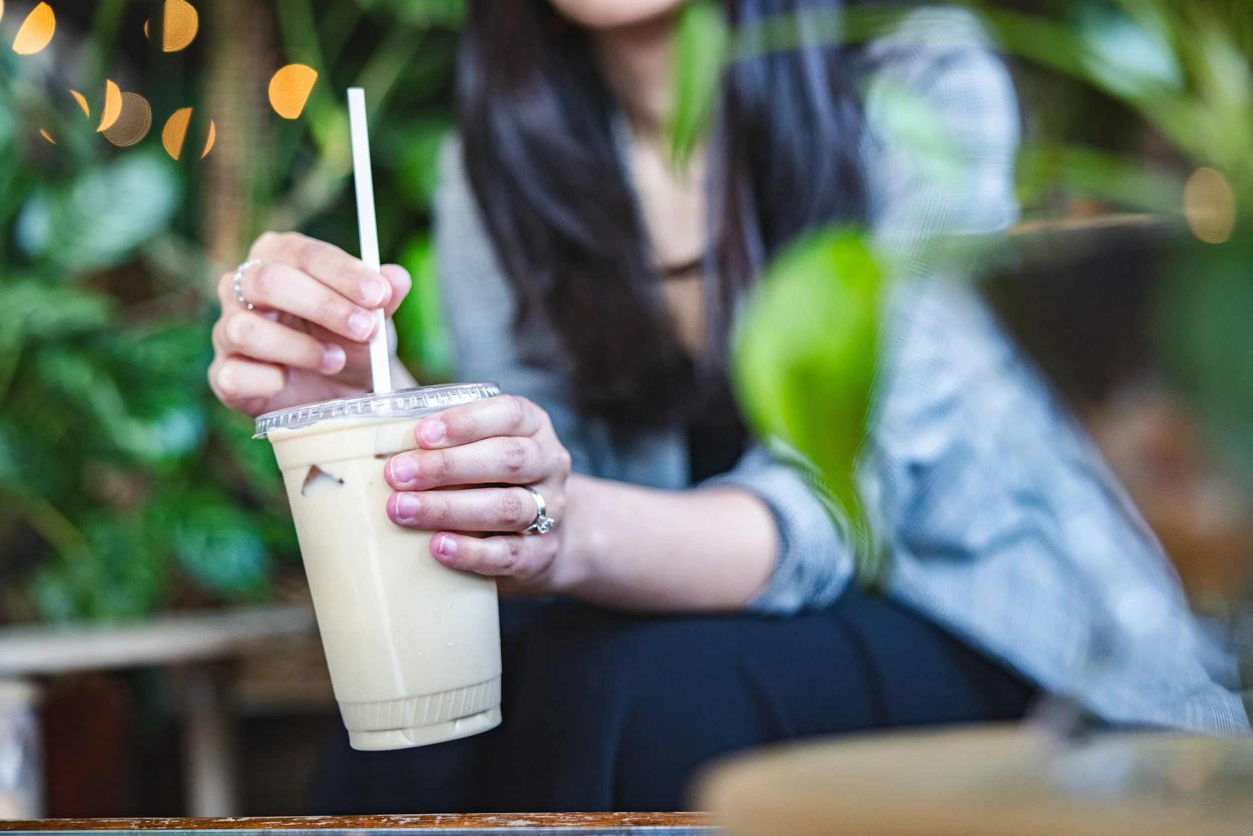 A person holding a plastic cup of iced coffee with a straw in a lush green environment