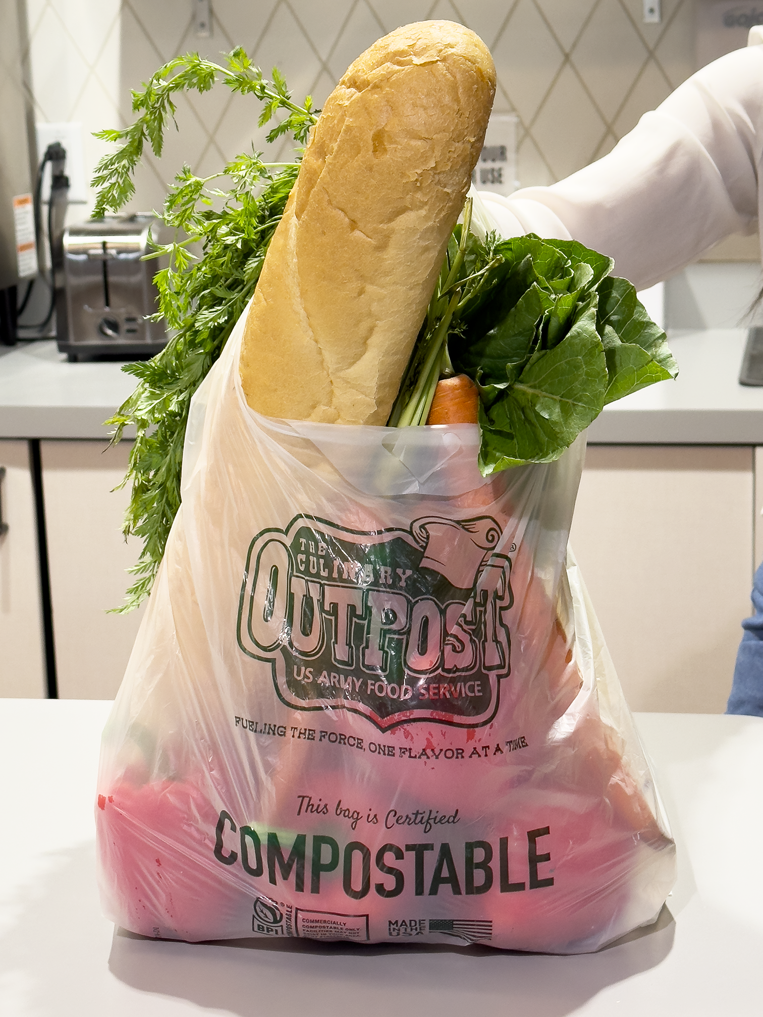 A plastic bag labeled 'Compostable' containing a loaf of bread, leafy greens, and a carrot, held by a person in a white shirt in a kitchen.