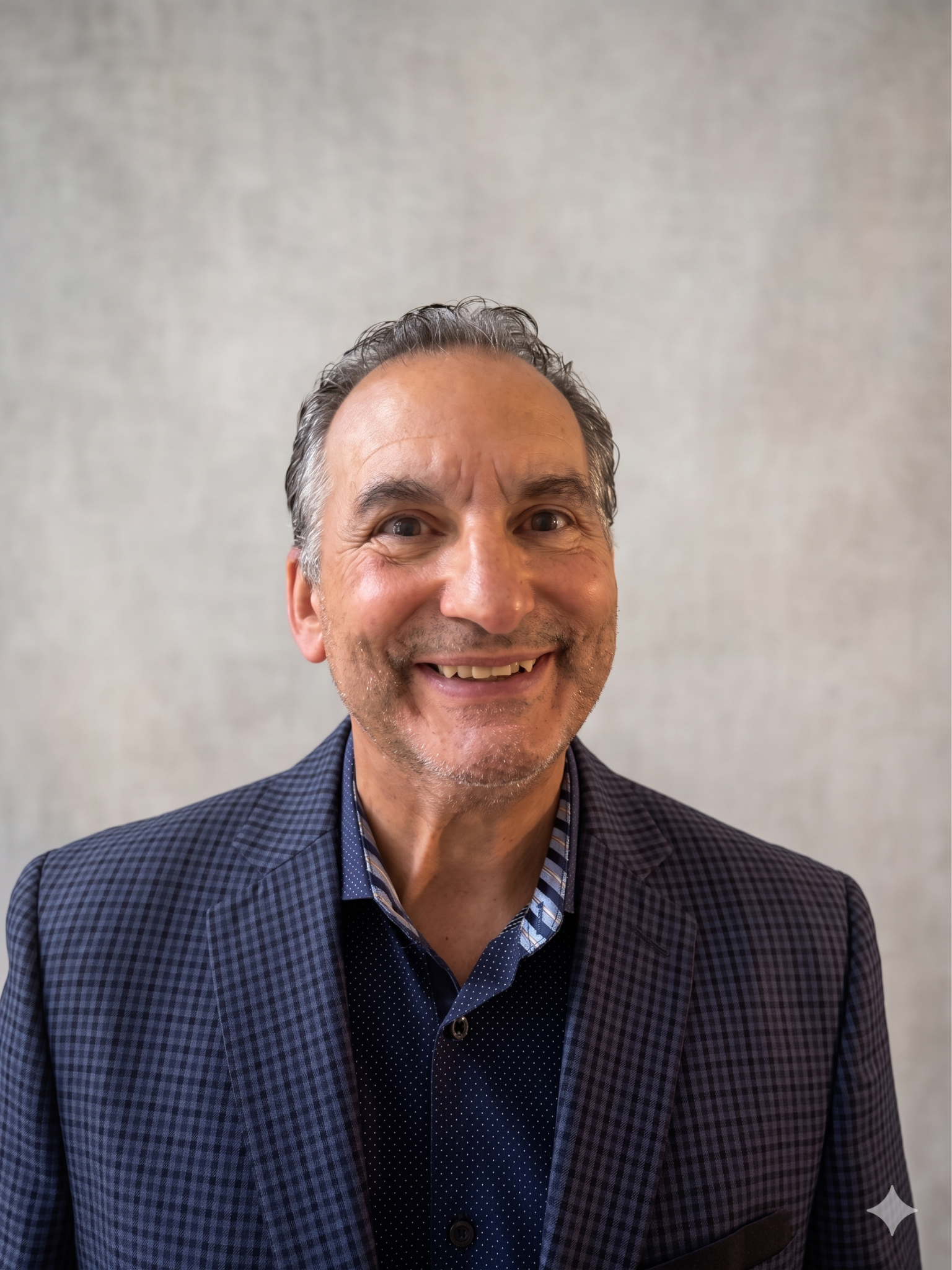 Close-up of a smiling middle-aged man with slicked-back hair, wearing a dark checkered blazer and a collared shirt with a patterned tie, standing against a plain light-colored wall.