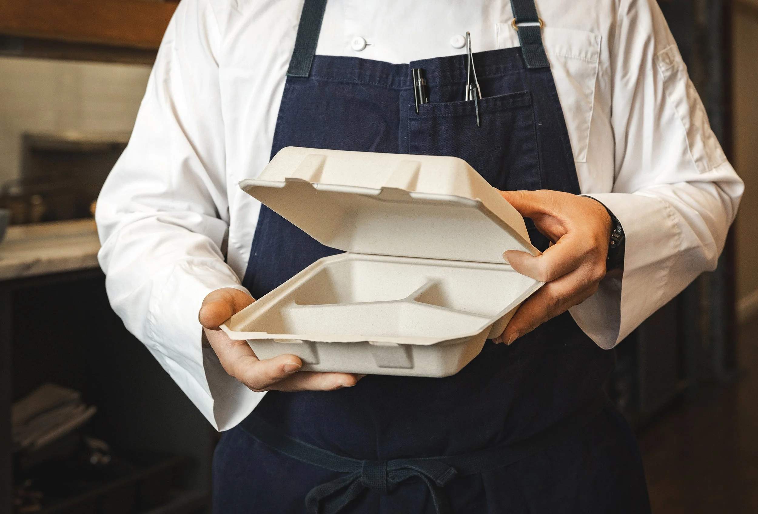 Person wearing a white shirt and dark apron holding a disposable takeout container with food inside.