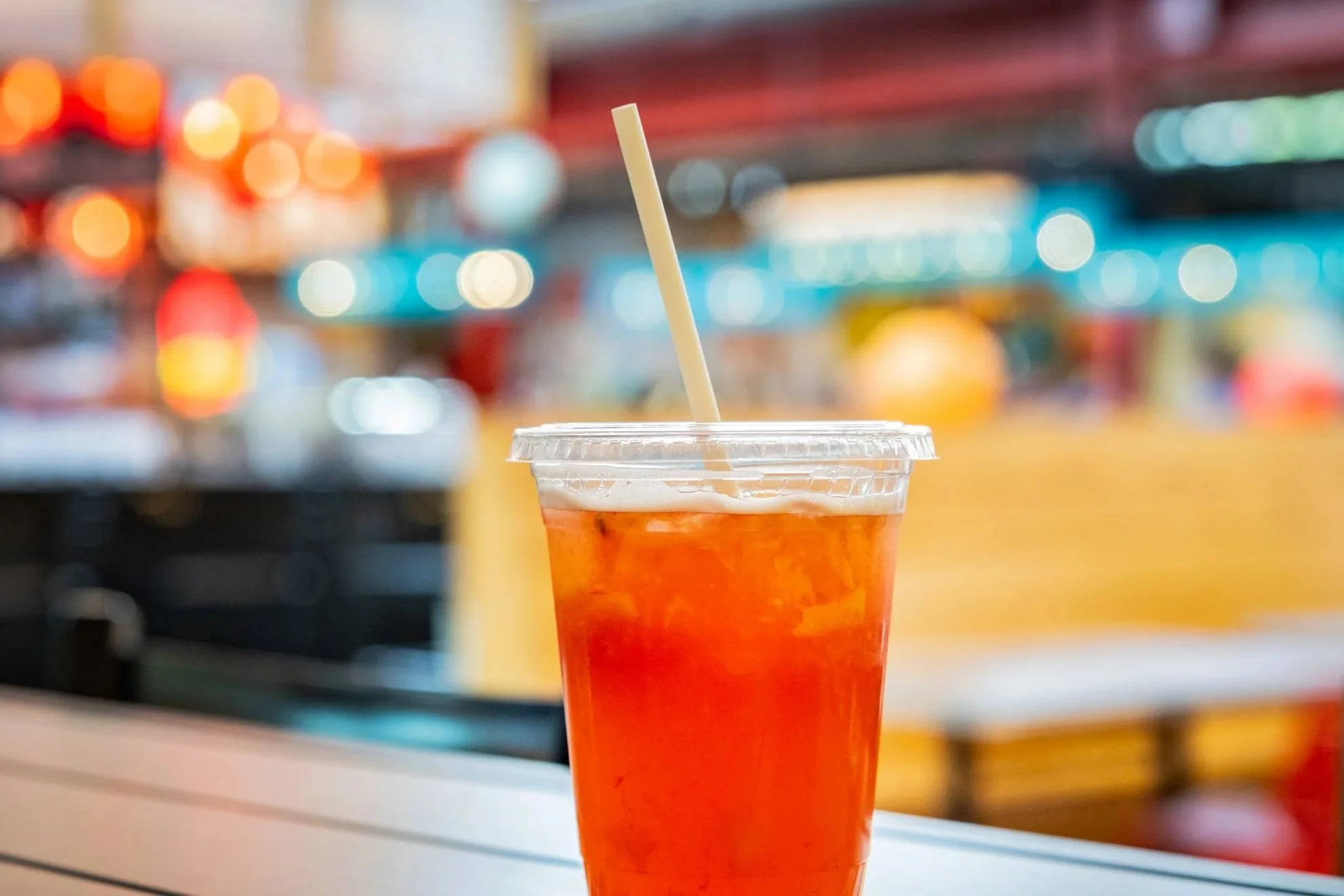A clear plastic cup filled with a red-orange beverage, topped with ice, and a white straw sticking out of the cup. The background is blurred with colorful lights and a store setting.