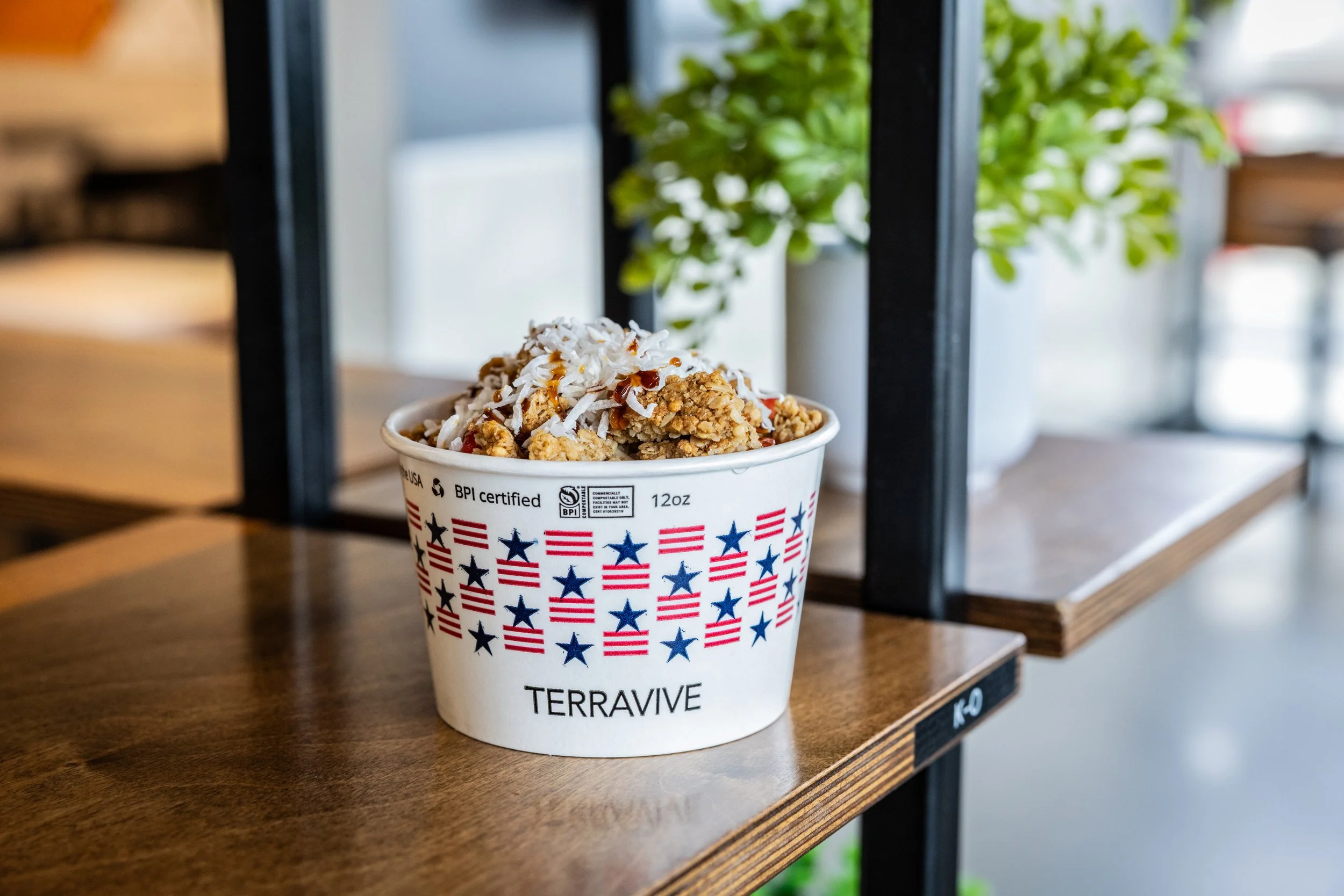 Cup of food with granola, coconut flakes, and drizzled sauce, placed on a wooden surface with a metal frame and greenery in the background.
