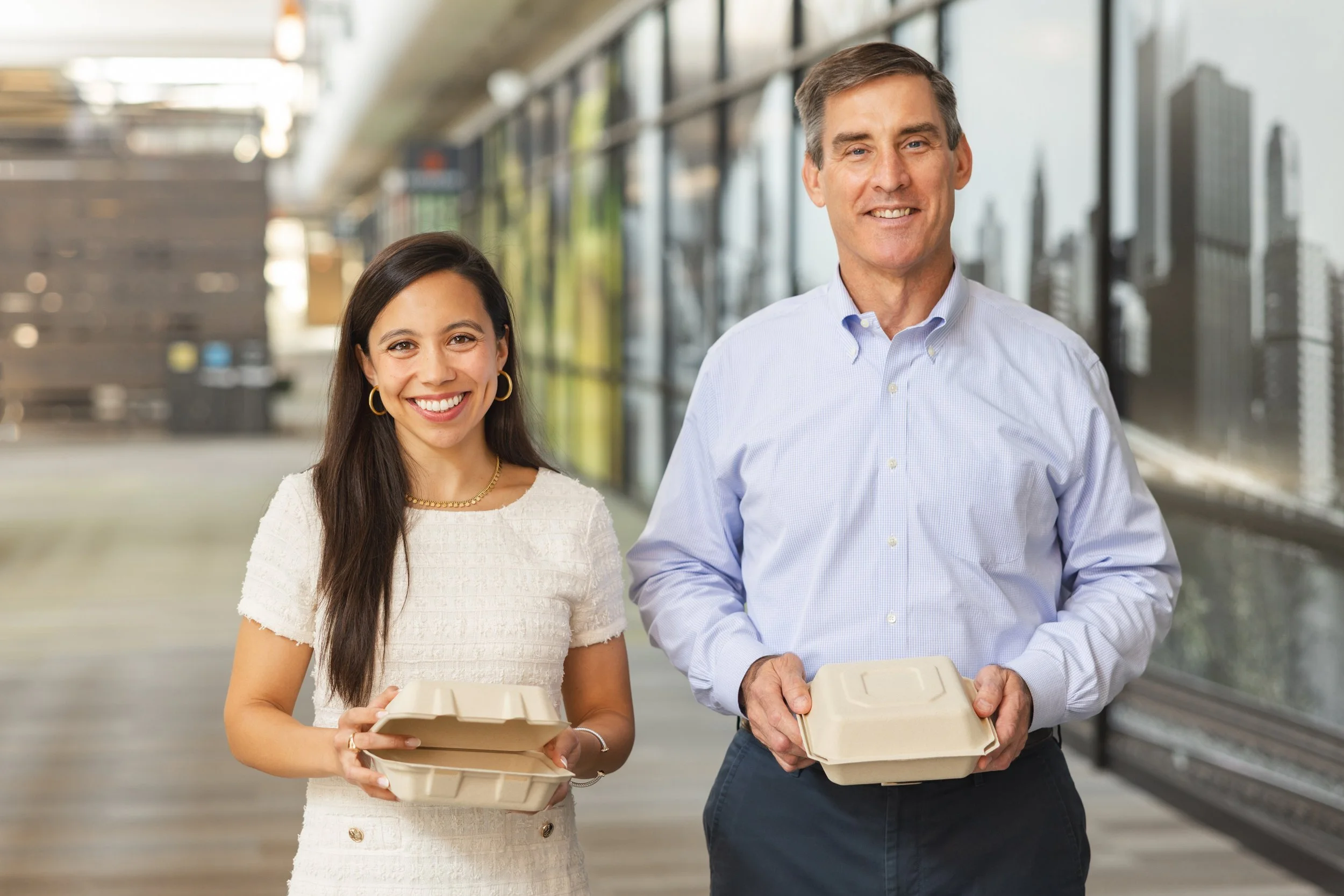 A young woman and an older man standing side by side outside, holding takeout food containers, smiling at the camera.