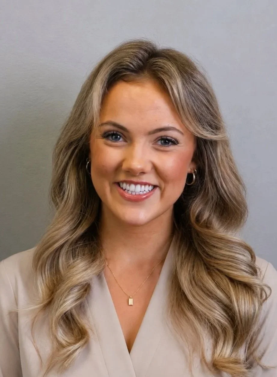 A smiling woman with long wavy blonde hair, wearing hoop earrings, a gold necklace, and a beige top, standing against a plain gray background.