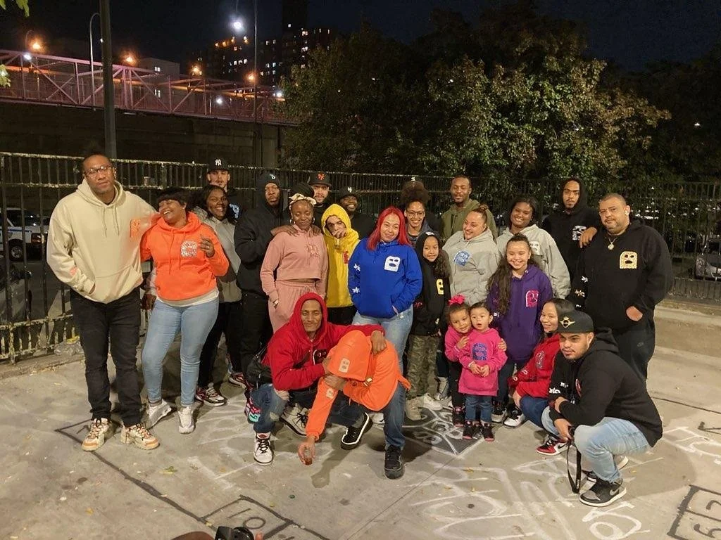 Group of adults and children gathered outdoors at night for a photo, with some wearing colorful hoodies and jackets, and a chalk drawing on the ground.
