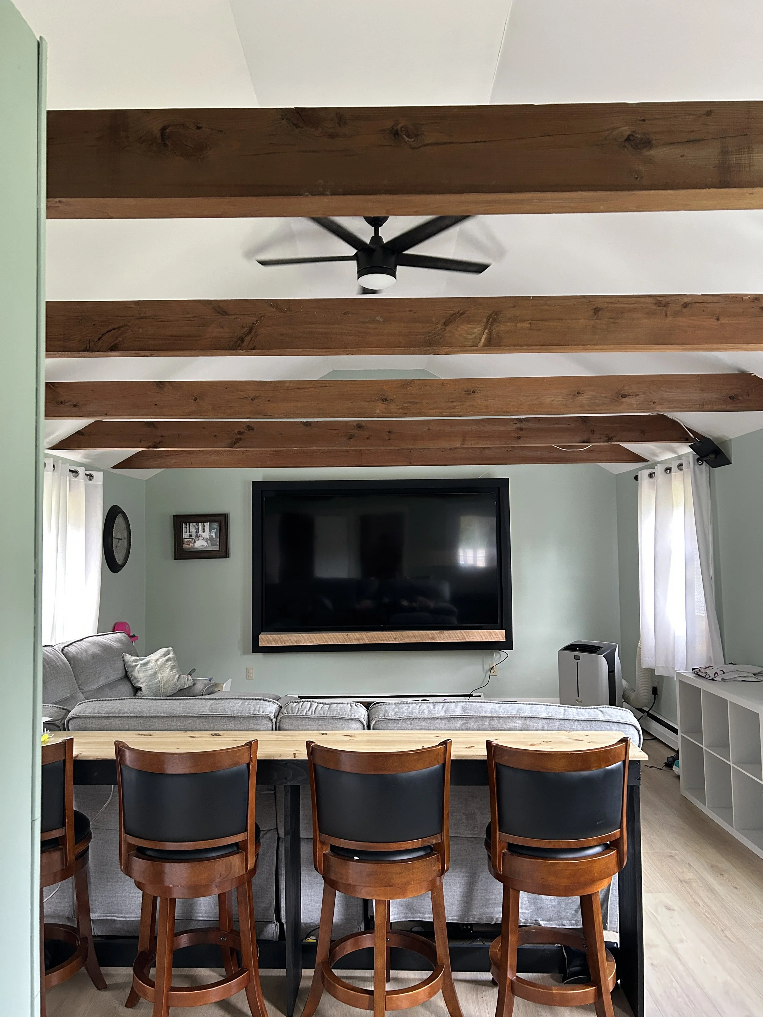 A living room with wooden beams ceiling, a wall mounted flat screen TV, a split heater on the floor, and a gray sofa with pillows. There are windows with white curtains, and a multicolored poster or picture frames on the wall.