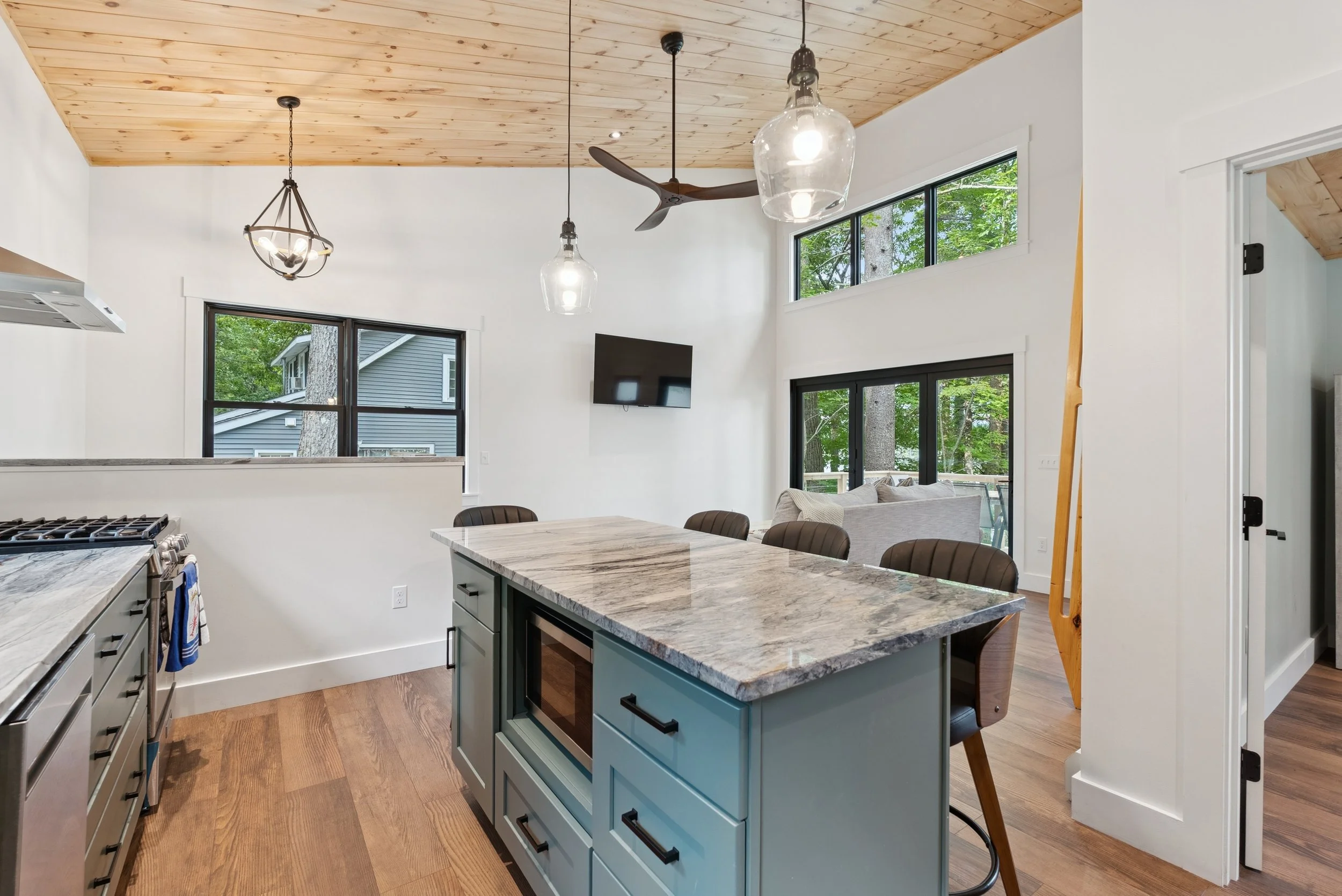 Modern kitchen with a marble island, wooden floors, large windows, and a wooden ceiling, with a dining area visible.