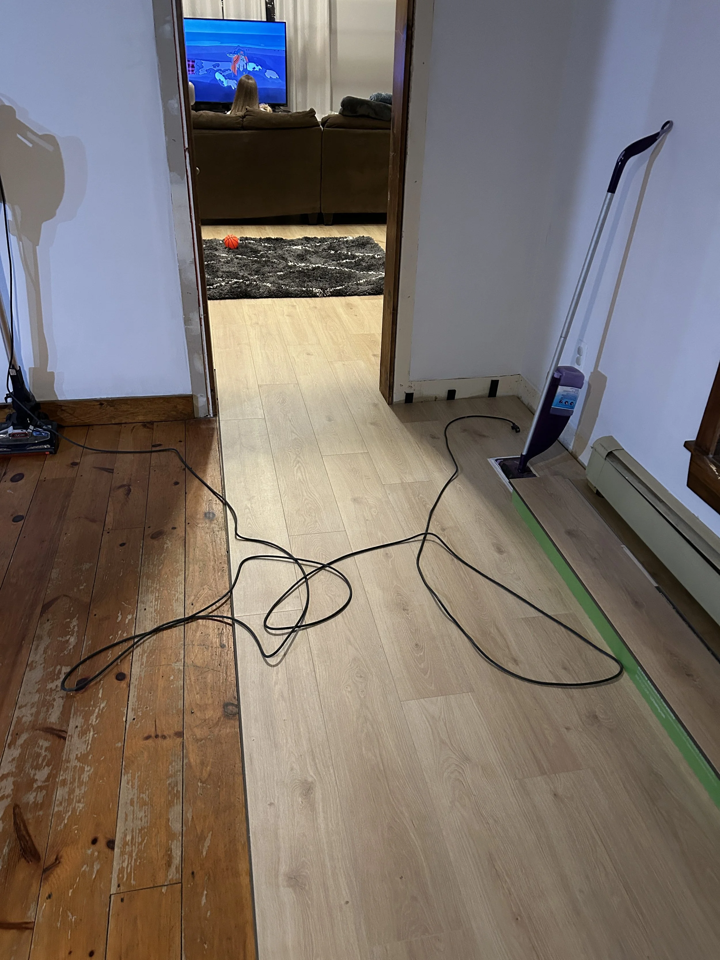 View of a living room seen through an open doorway. In the living room, there is a person watching a cartoon on a large TV. On the floor near the doorway, there are some black cords, a mop with a purple handle, and a box or toolbox. The room has two-
