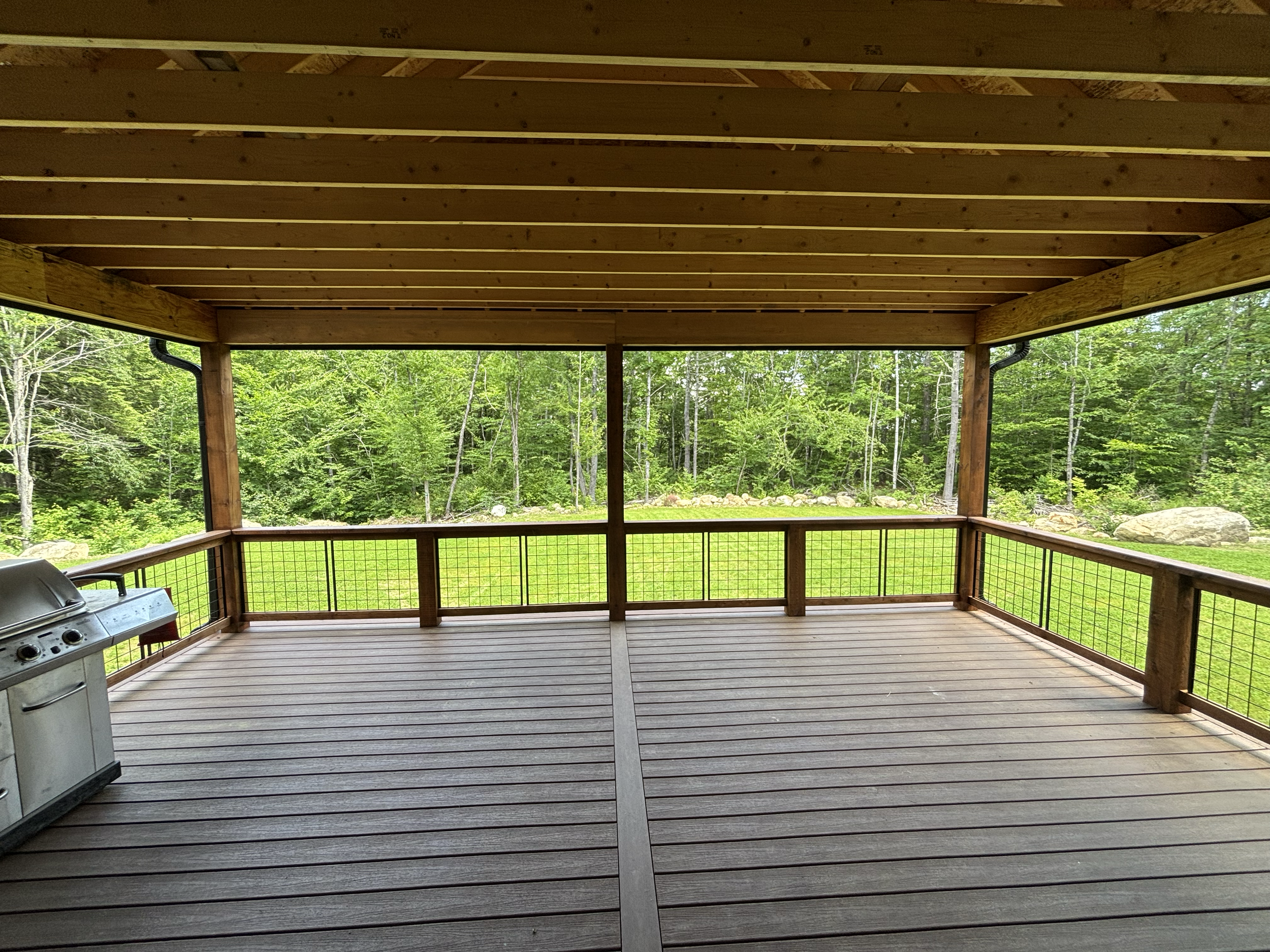 This image shows a covered wooden deck with a view of green trees and rocks in the background. There is a, small BBQ grill in the lower left corner and a separating wire mesh along the edges of the deck.