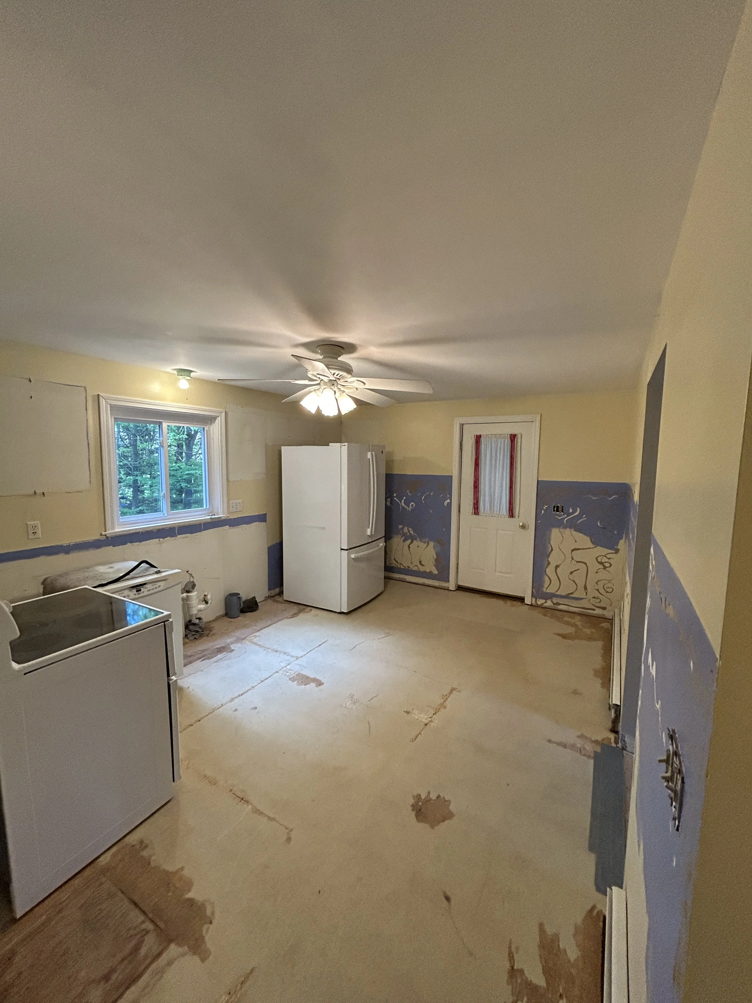A kitchen under renovation with partially removed flooring and painted walls. The room has a window, a ceiling fan with lights, a refrigerator, and a washing machine all ready for removal. 