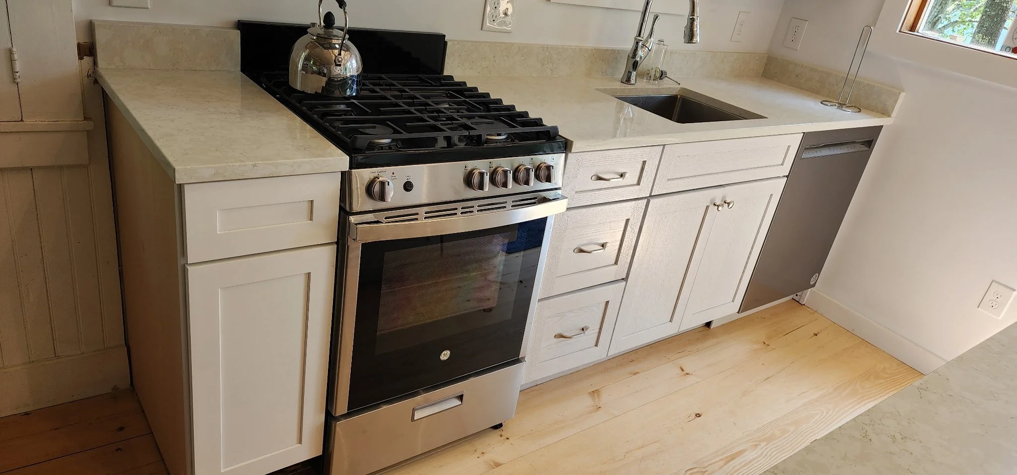 A kitchen countertop with a stovetop, kettle, sink, and a dishwasher, with white cabinets below and a window to the right.
