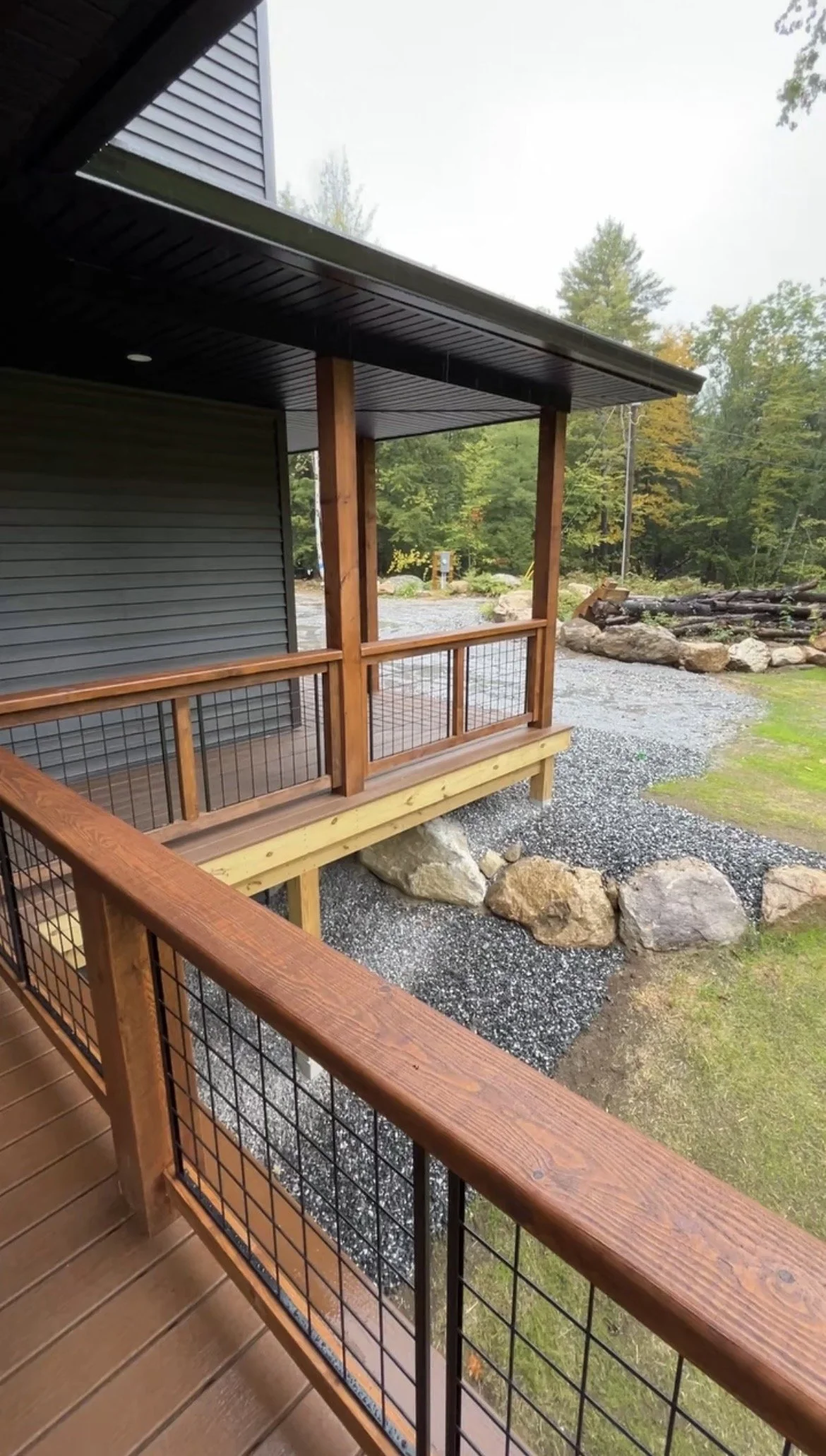 Wooden porch railing with metal grid, view of gravel yard with rocks and trees in the background.