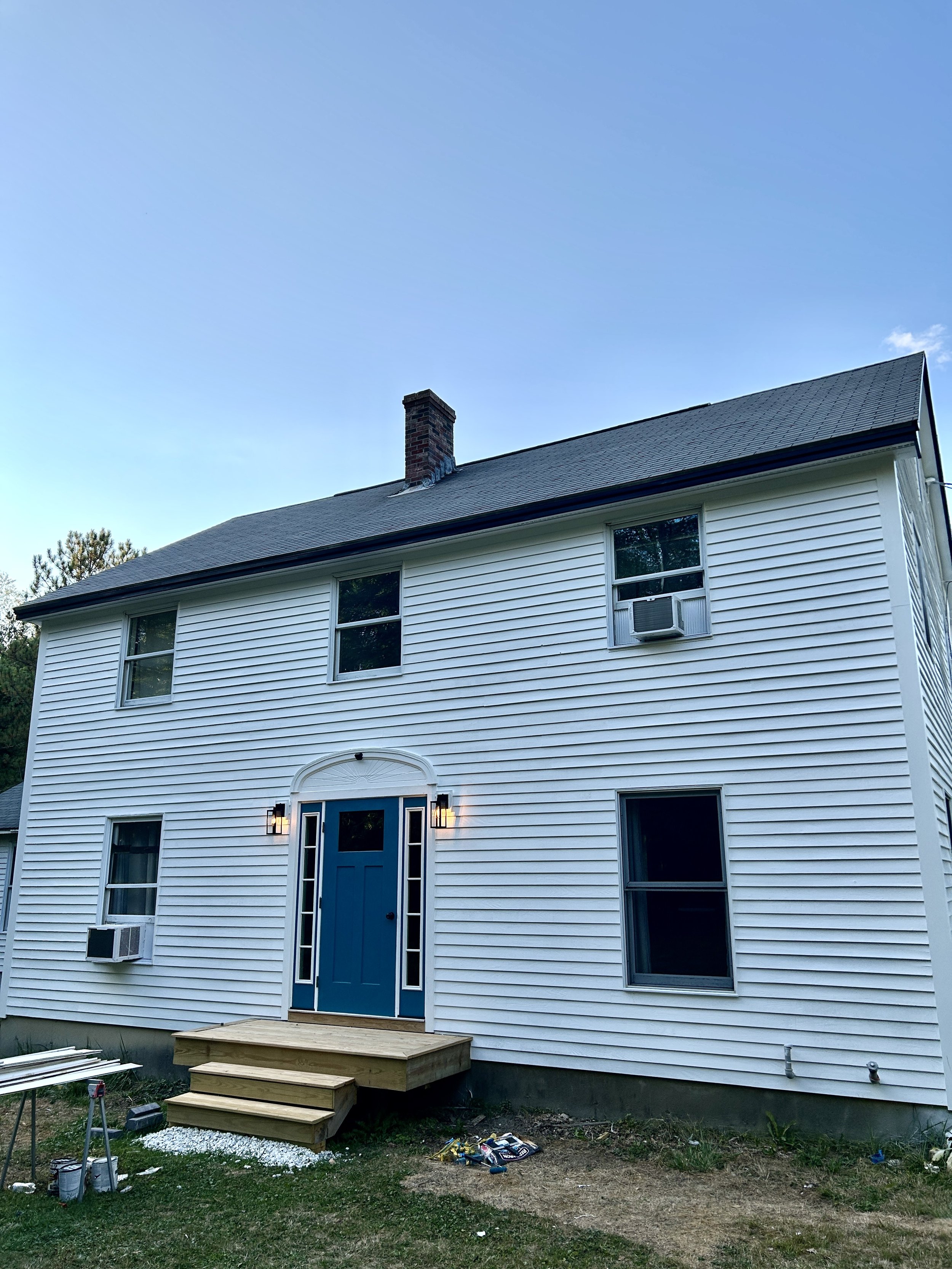 The back of a white house under construction with a blue door, multiple windows, and a small wooden porch with stairs. There are construction materials and tools on the ground nearby.