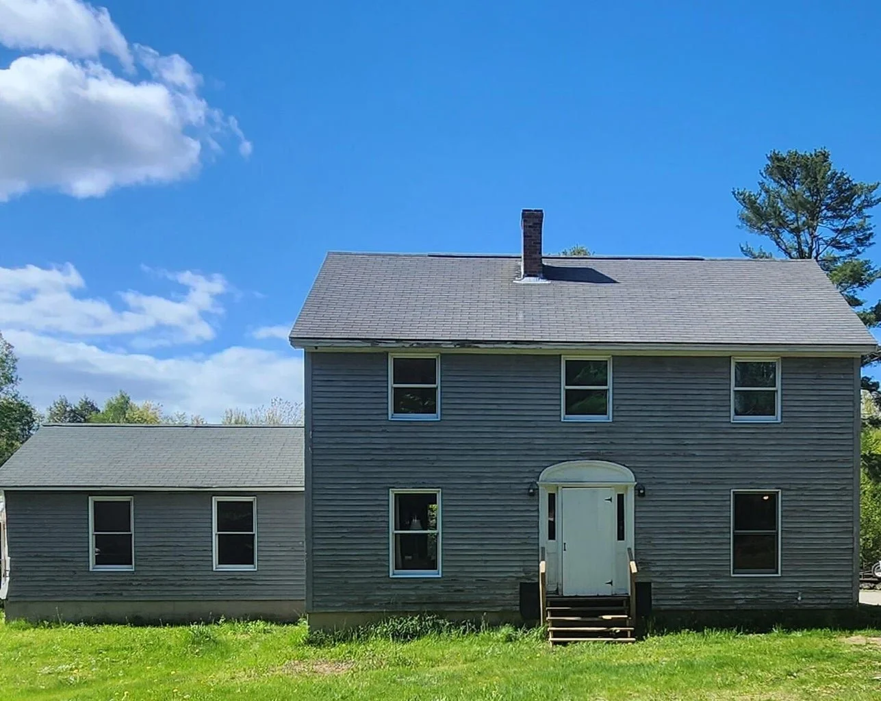 A two-story gray house with four windows on the upper floor, three windows on the lower floor, and a white front door with stairs and a small porch. The house has a chimney and is surrounded by a grassy yard, with a blue sky and some clouds overhead.