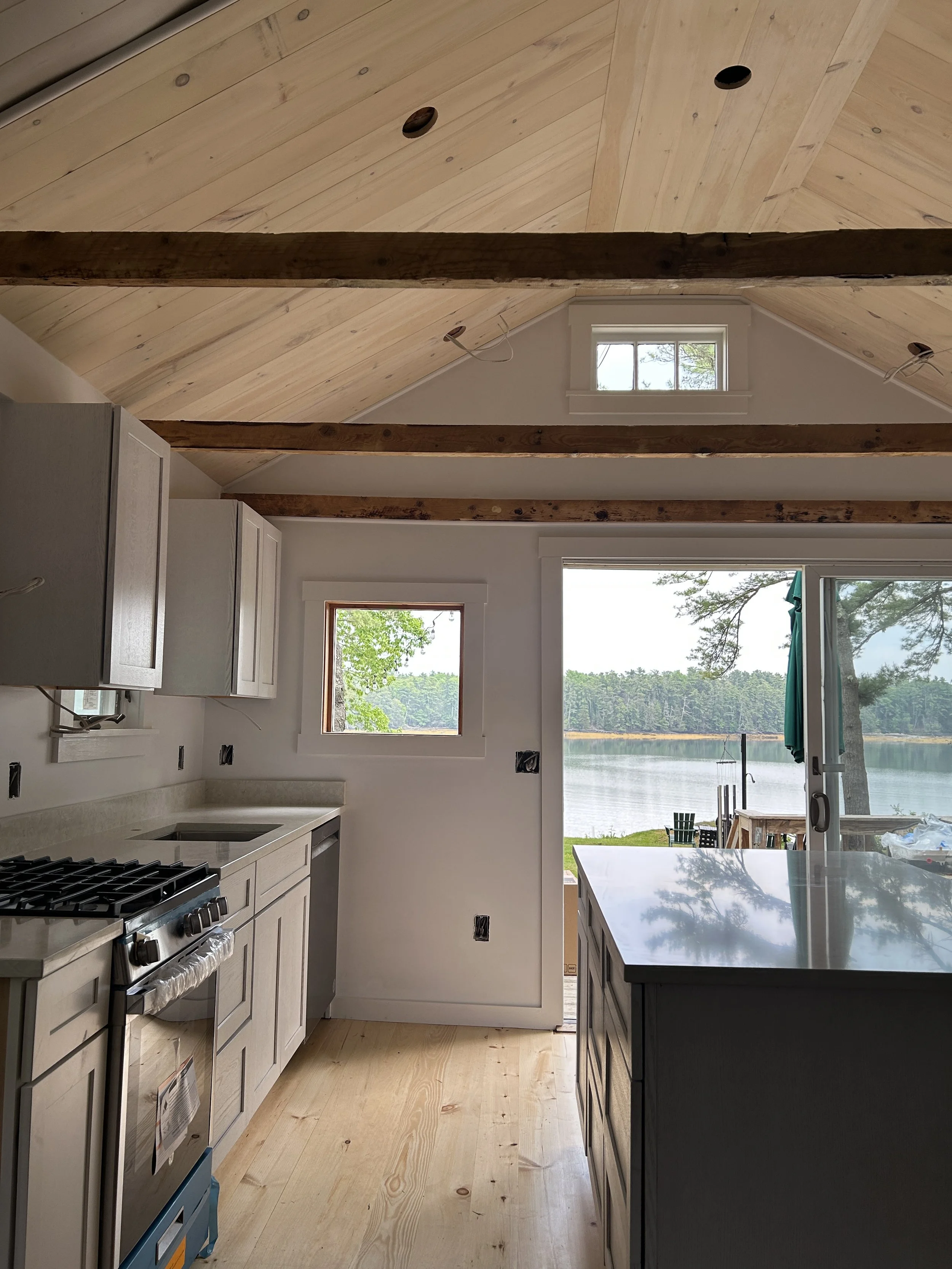 A kitchen with light wood cabinets, a white island with a marble top, a stainless steel stove, white walls, and large windows showing a lake and trees outside.