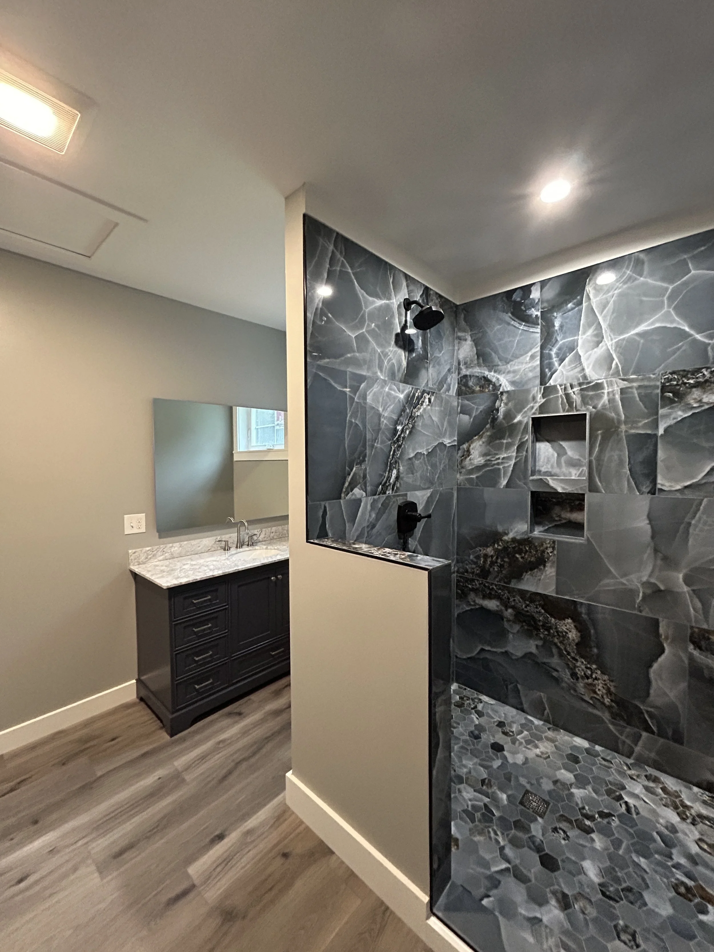 Bathroom with a walk-in shower featuring dark gray marble tiles, a black showerhead, and a small built-in shelf. Adjacent to the shower, there is a vanity with a sink, dark cabinetry, and a large mirror. The bathroom has light-colored walls, a window
