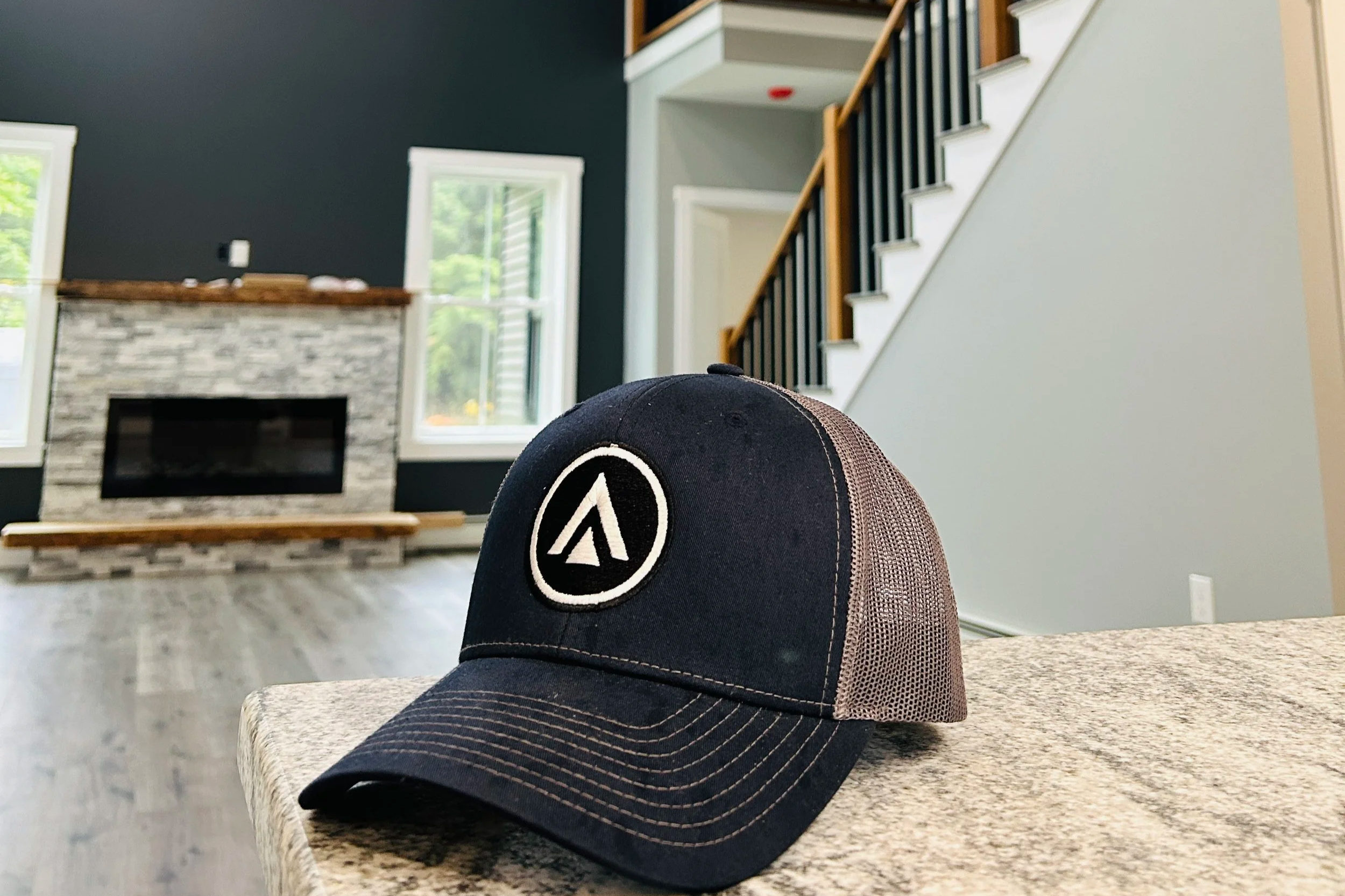 A black and beige trucker cap with a circular logo on the front, placed on a granite countertop in a living room with a fireplace, large windows, and a staircase.