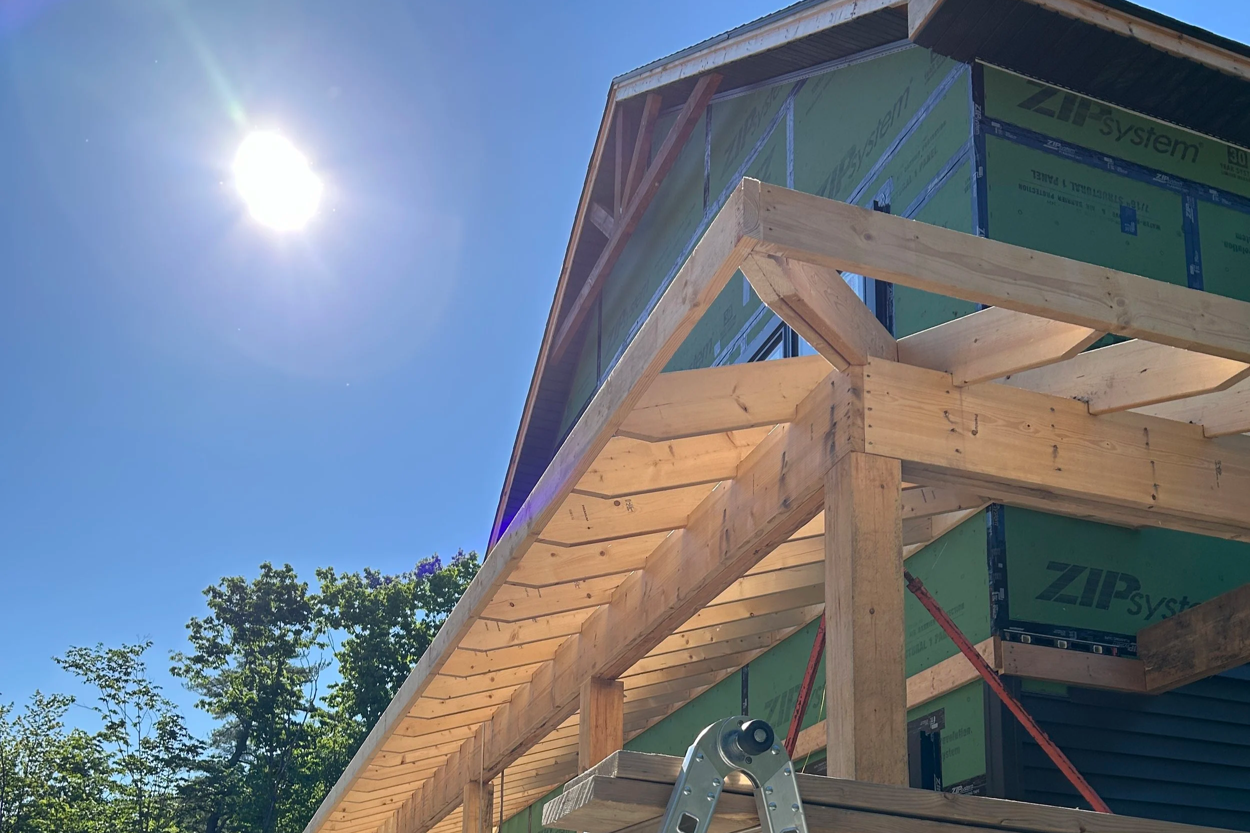 Construction of a wooden building framework with a blue sky, sun, and trees in the background.