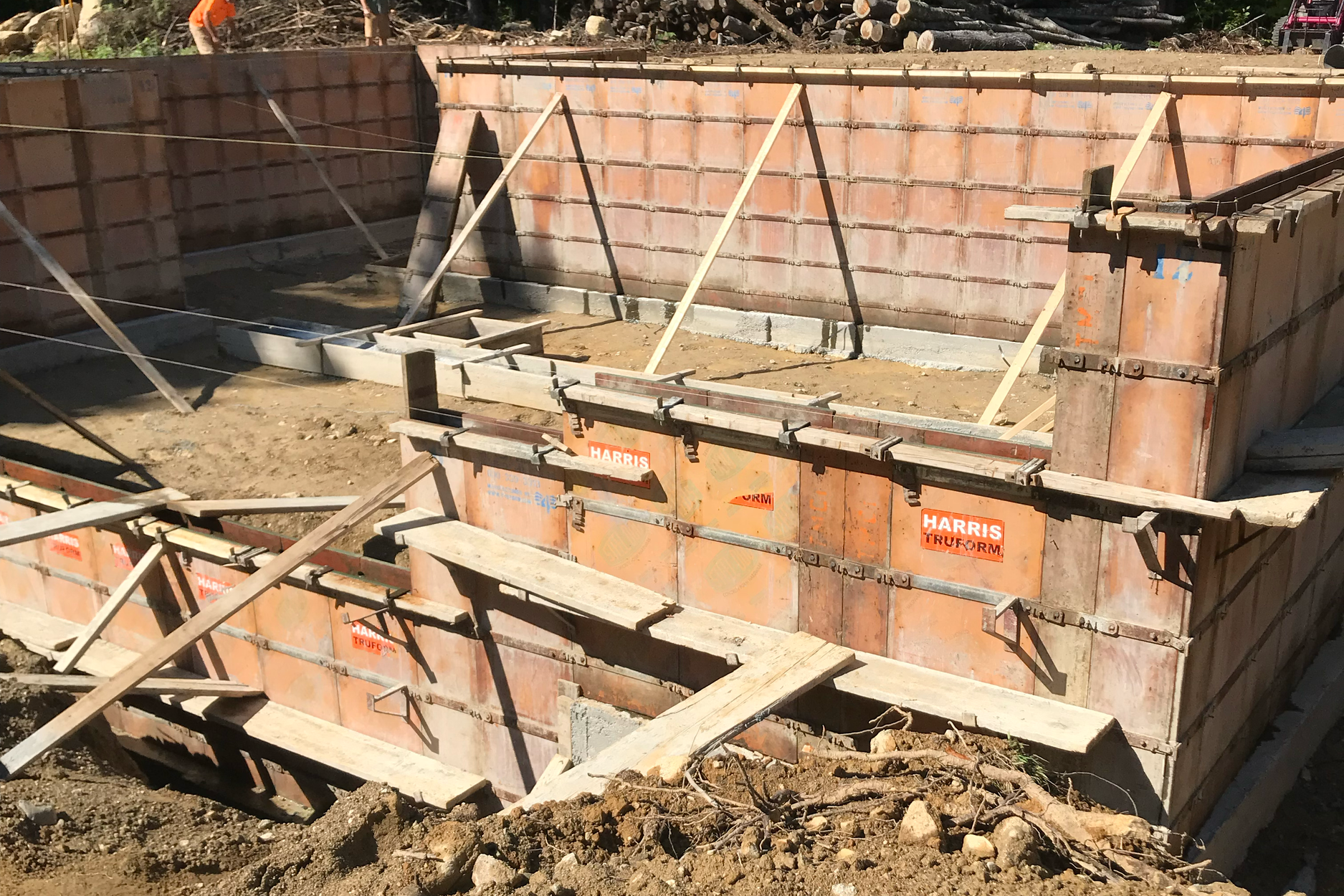 Concrete foundation formwork at a construction site with wooden supports, metal clamps, and orange Harris Tru-Form panels.