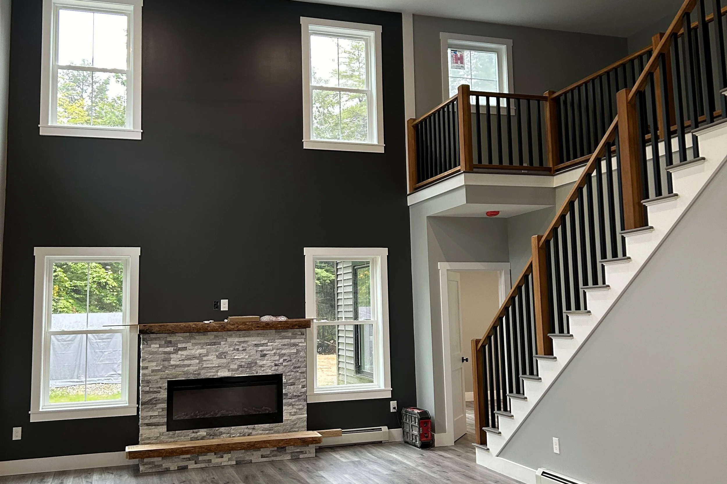 Interior of a modern living room featuring high ceilings, large windows, a stone fireplace, and a staircase with wooden and black railing.