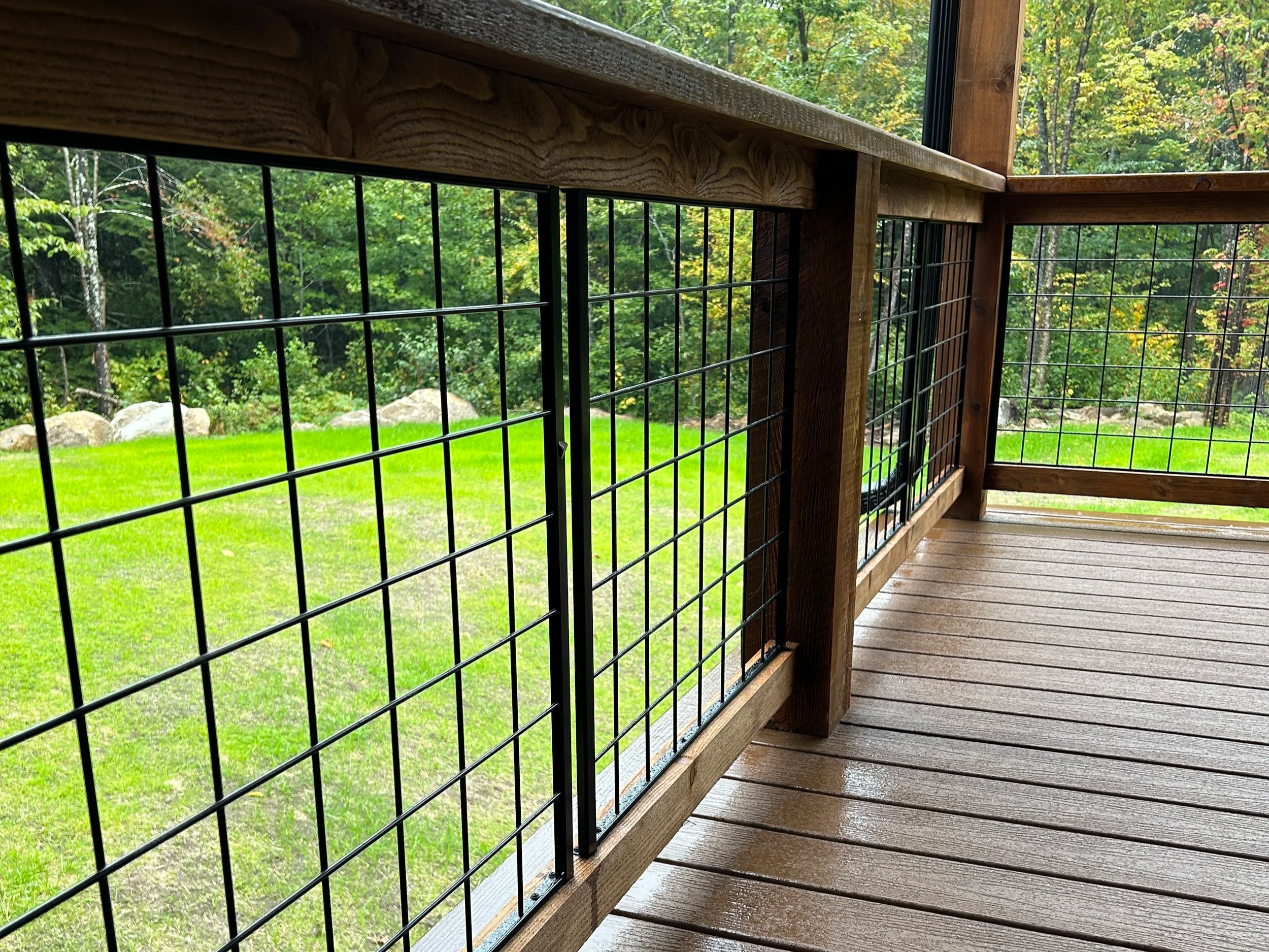 Close-up of a wooden deck railing with black wire grid fencing, overlooking a lush green yard and forest.