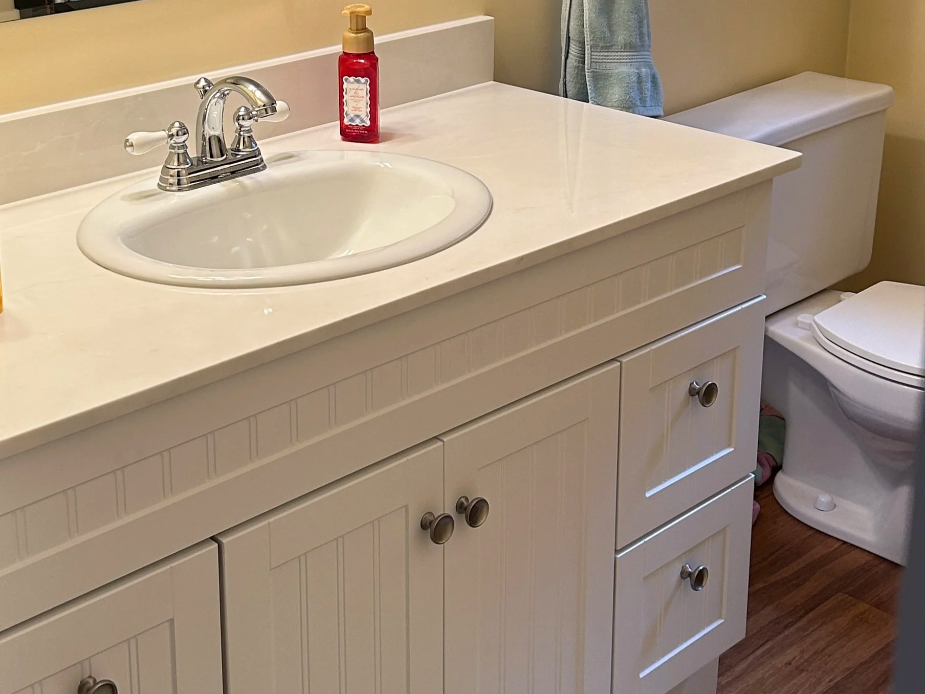 Bathroom vanity with a white sink, soap dispenser, and a cabinet below, next to a toilet with a top cover, a towel hanging on the wall, and hardwood flooring.