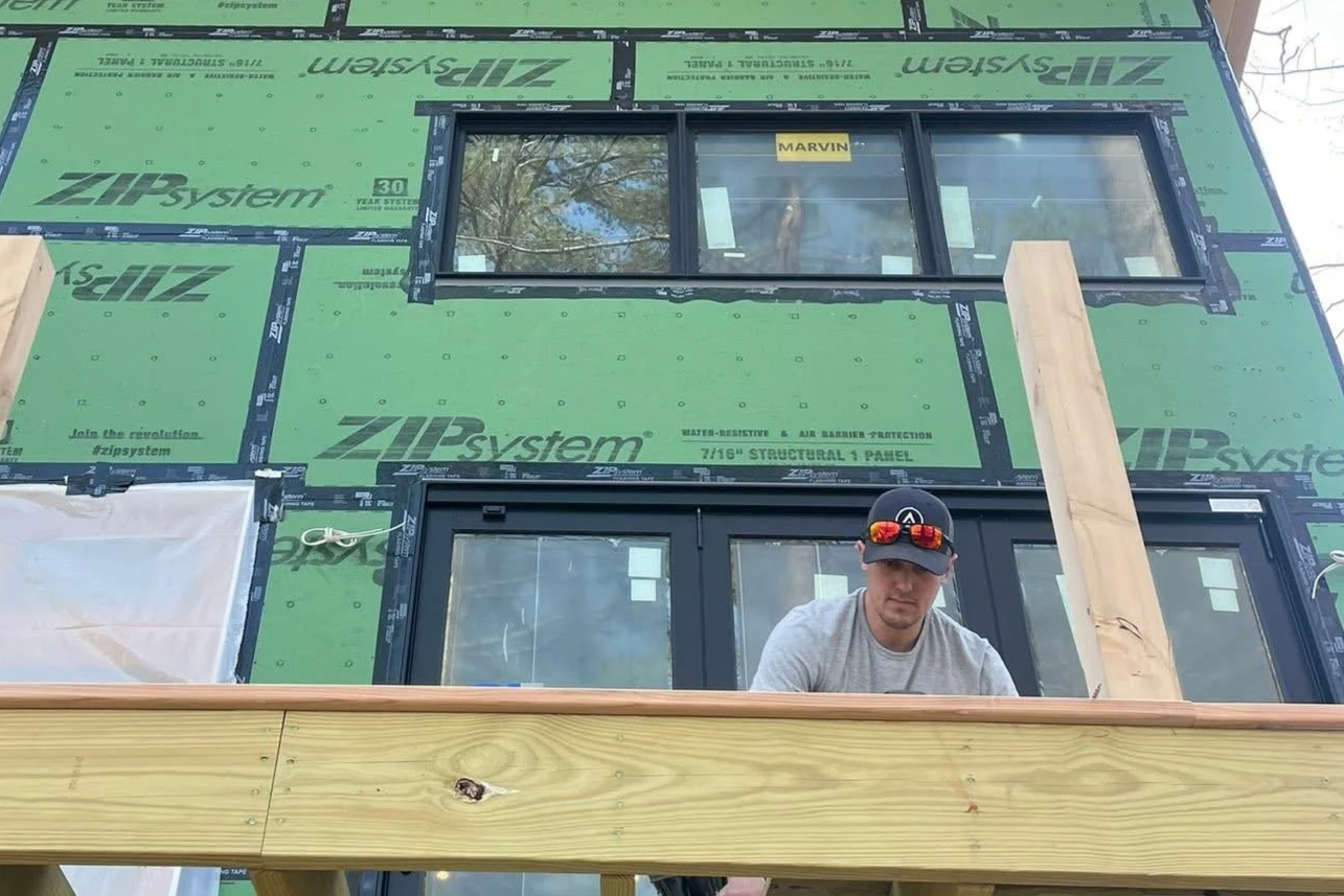 Avex Custom Homes owner Andrew is working on the exterior construction of a building, with green ZIP System sheathing and large window frames visible.