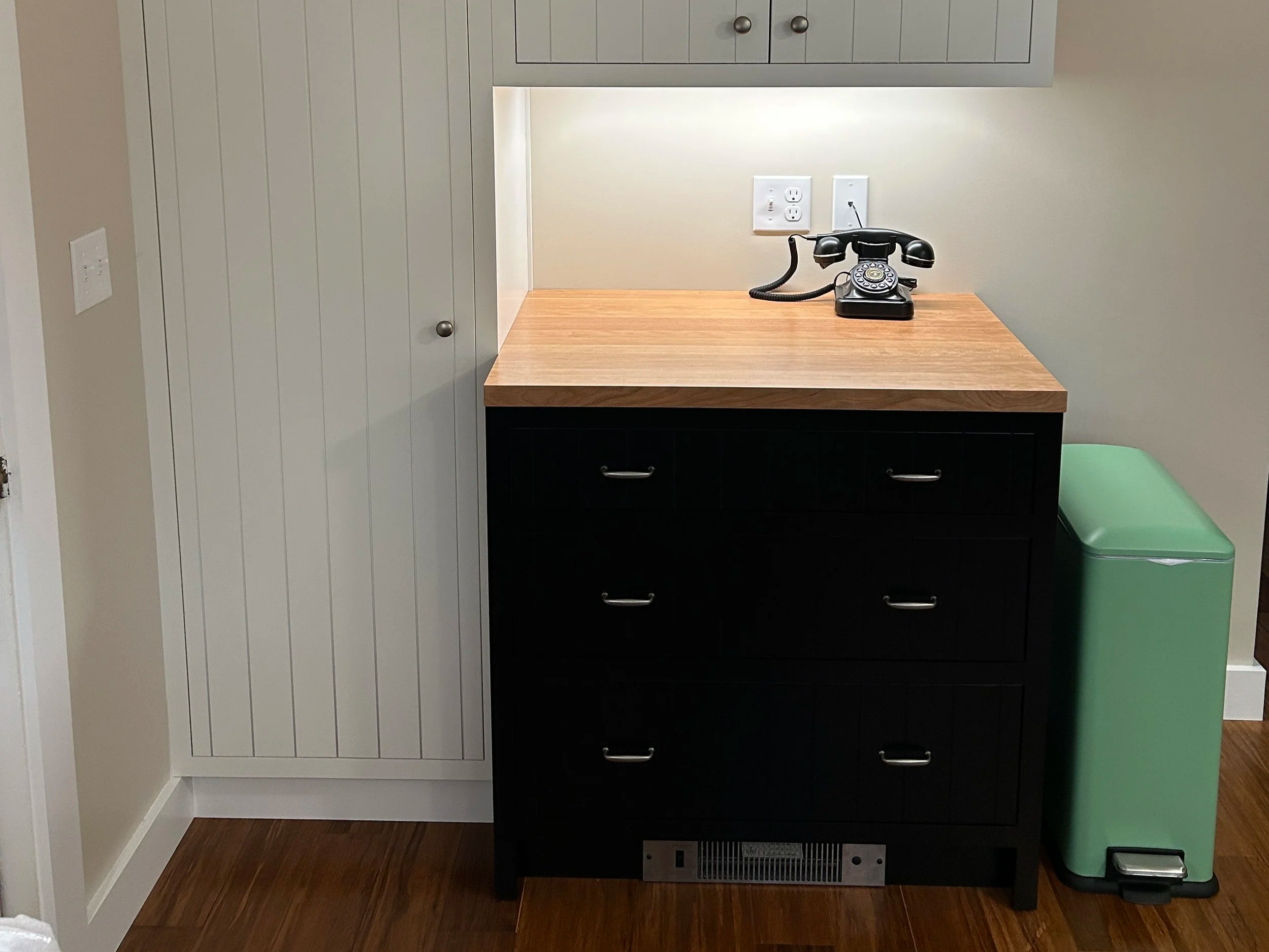A vintage black rotary dial telephone on a wood cabinet, with a white wall and electrical outlets behind it. There is a green trash bin next to the cabinet.