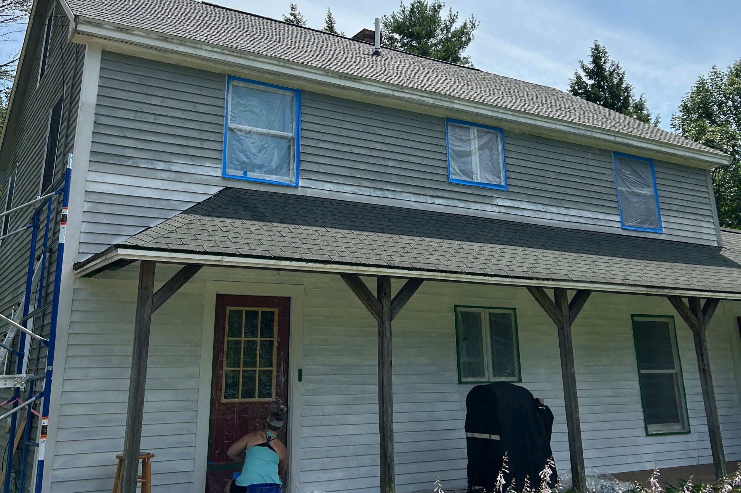 Back of a house with gray siding, under renovation, with three windows covered in protective plastic and blue painter's tape, and a scaffold on the left side. Two people are working near the door, one bending down and the other standing.
