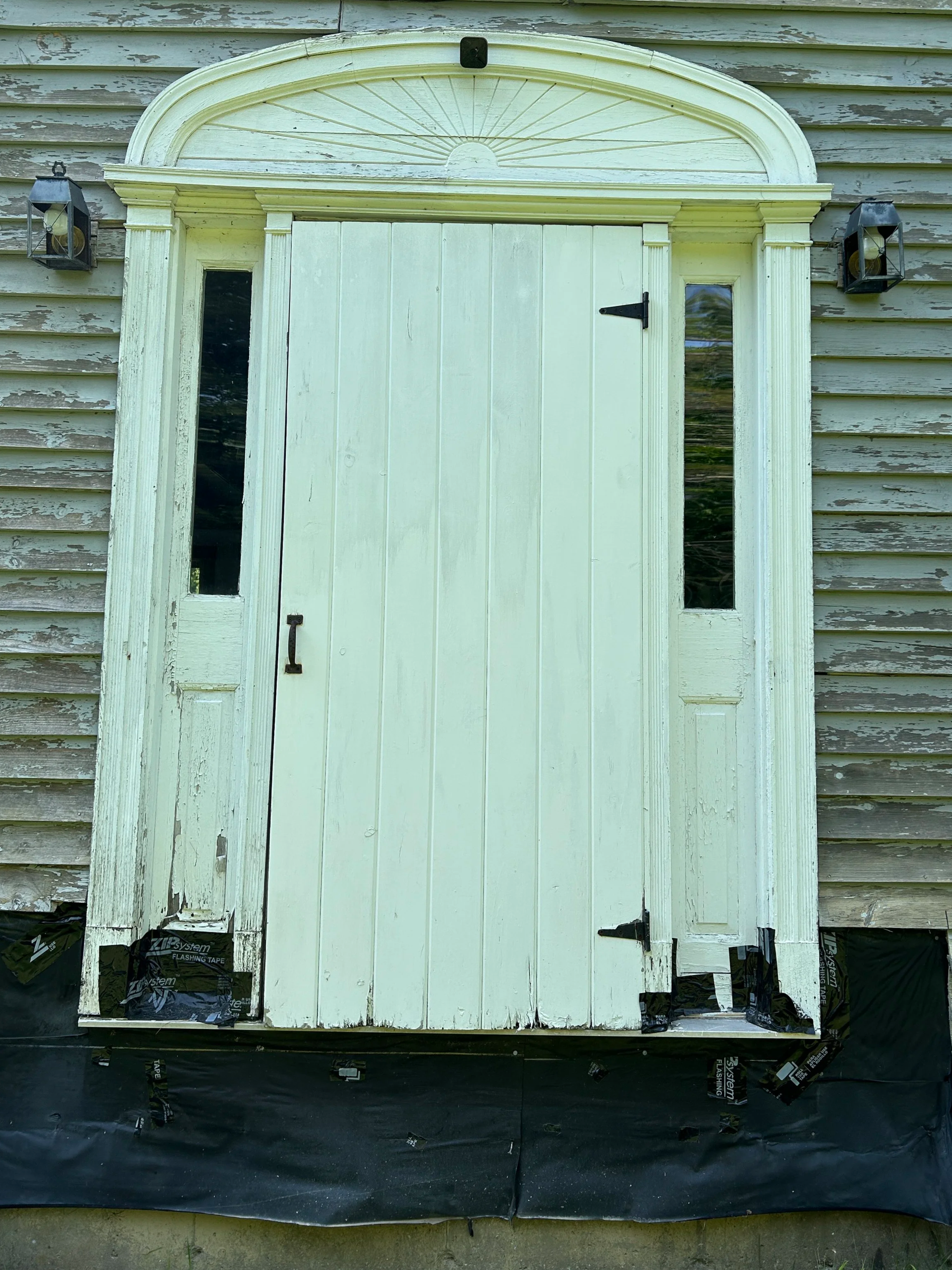 A white barn door with vertical panels, flanked by two narrow window panes, mounted on an aged, weathered wooden wall. Two black outdoor lanterns are attached on either side of the door, and partial black flashing tape is visible at the bottom.