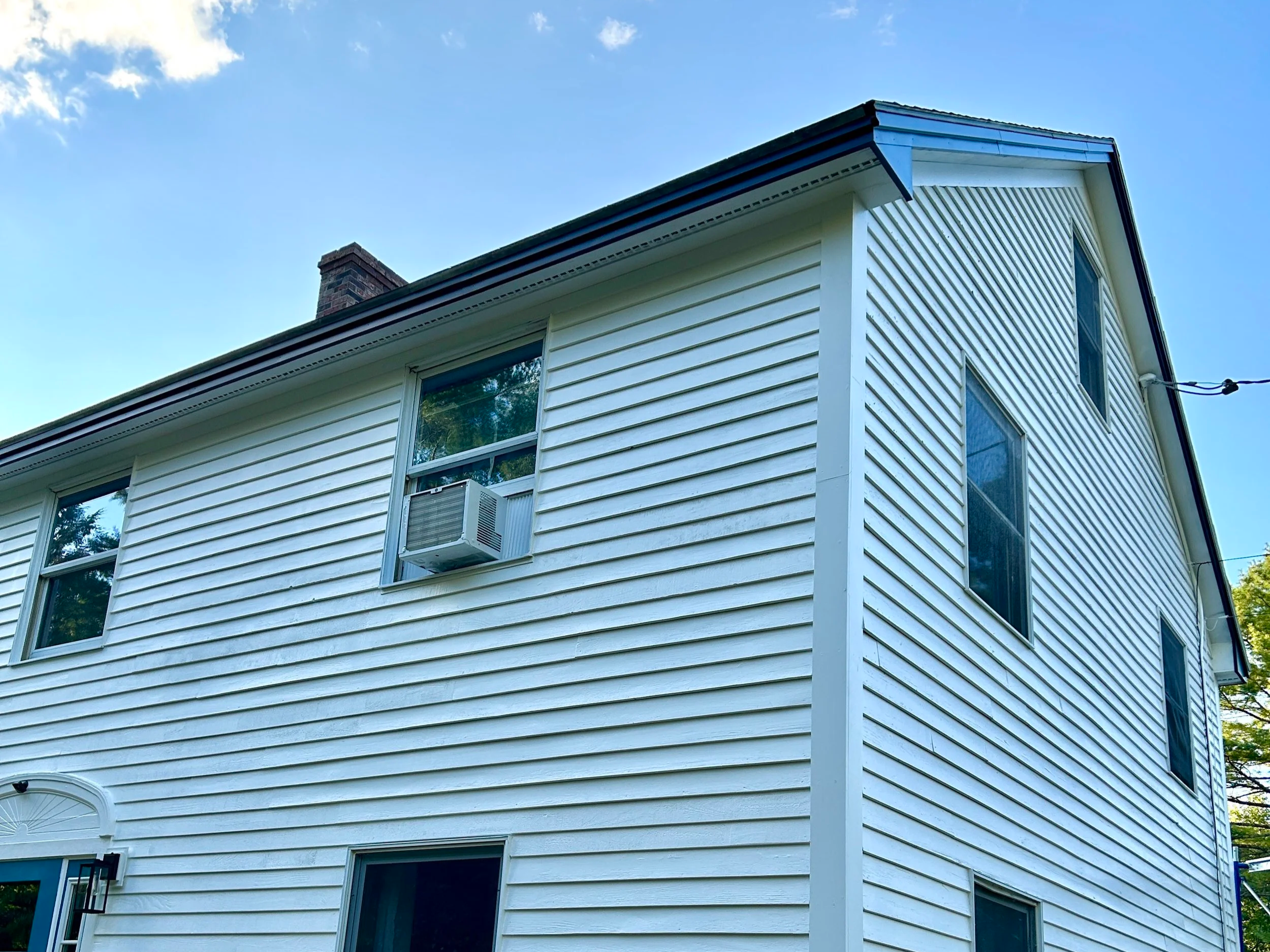 A white house with vinyl siding, multiple windows, a small air conditioning unit in one window, and a brick chimney on the roof, under a partly cloudy blue sky.