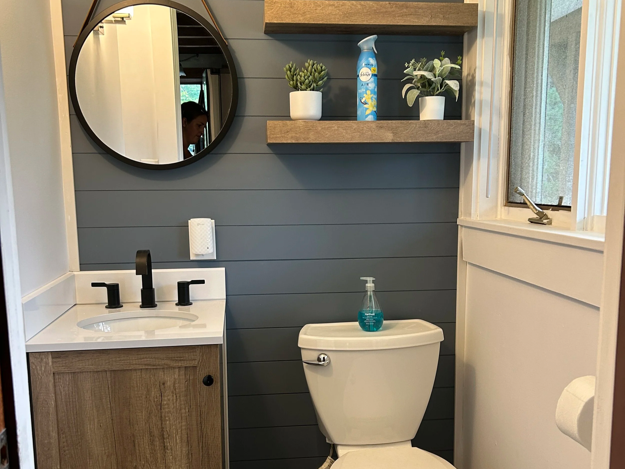 A bathroom with a modern design featuring a white sink with black fixtures, wood and gray wall panels, a black round mirror, two white potted plants on a wooden shelf, a white toilet with a bottle of hand soap on top, a window with a mesh screen, and