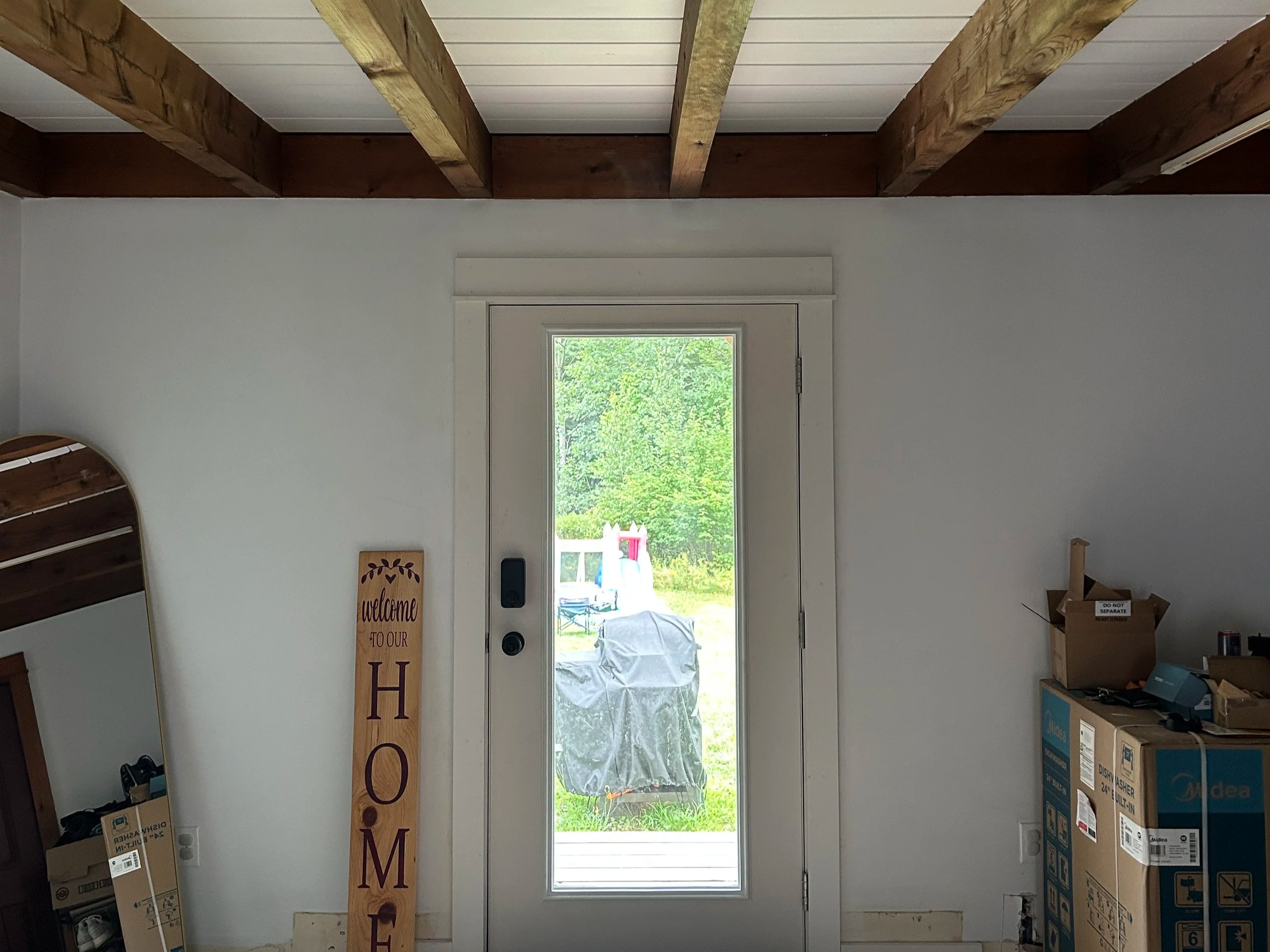 Interior of a room with white walls and a window showing a green landscape outside; wooden ceiling beams and cluttered items including boxes and a welcome sign.