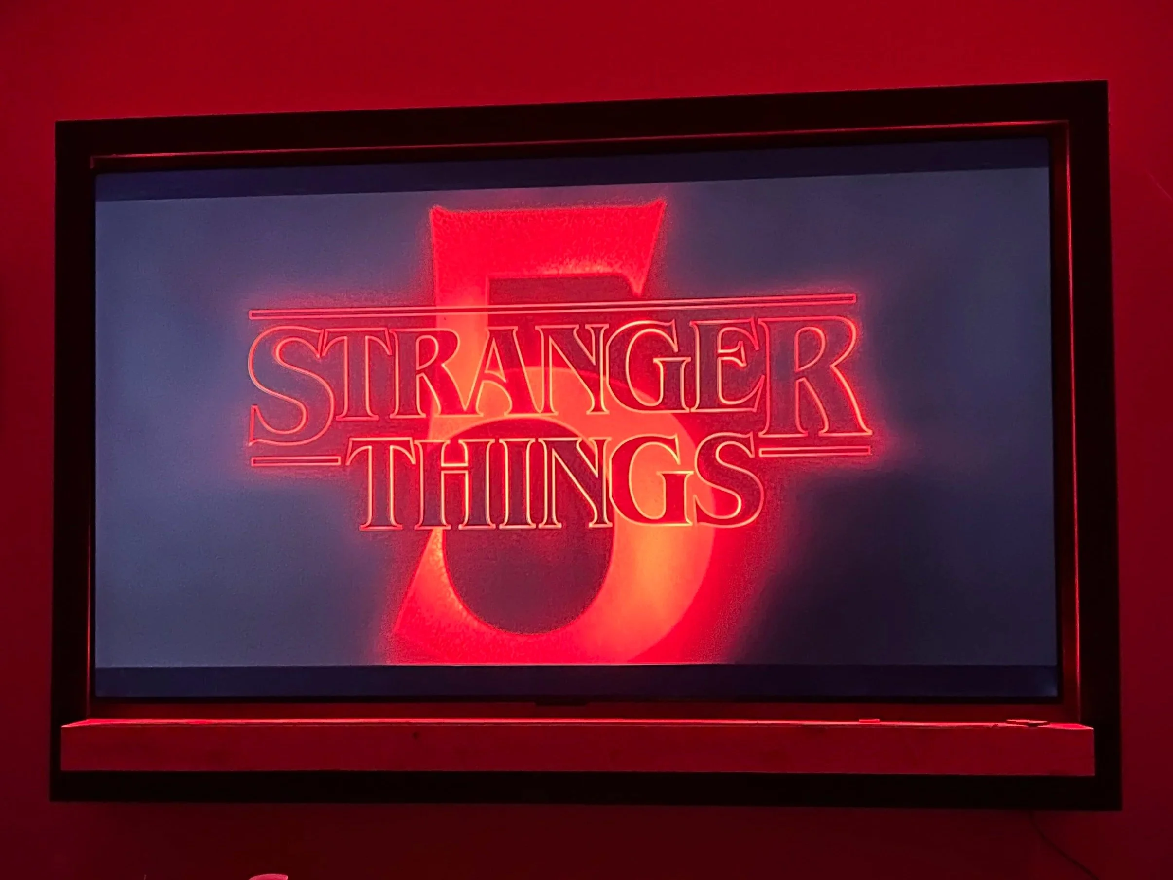 A television with the words "Stranger Things" glowing in red, framed in black on a red led light lit background.