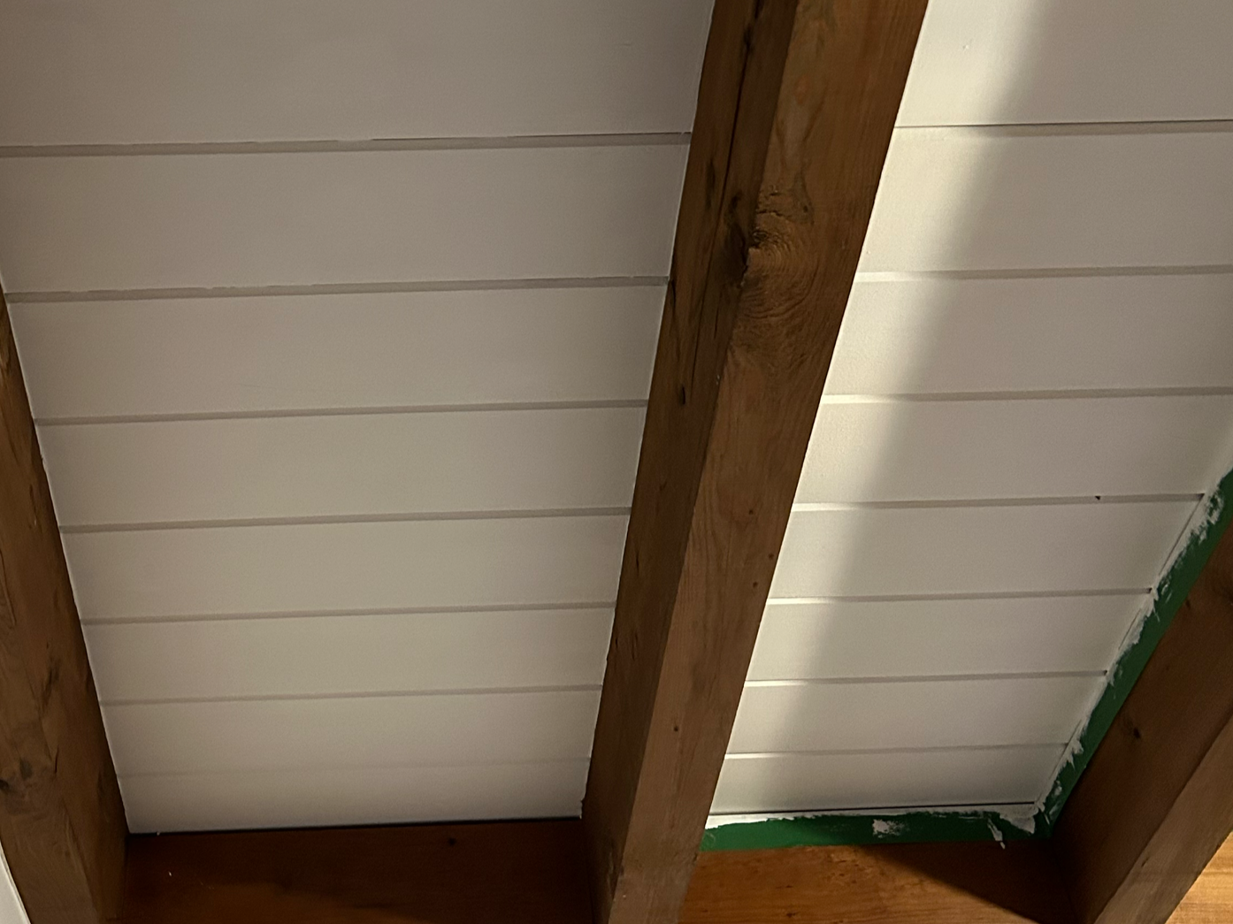 Ceiling with wooden beams and white paneling, with some green painter's tape and unfinished edges.