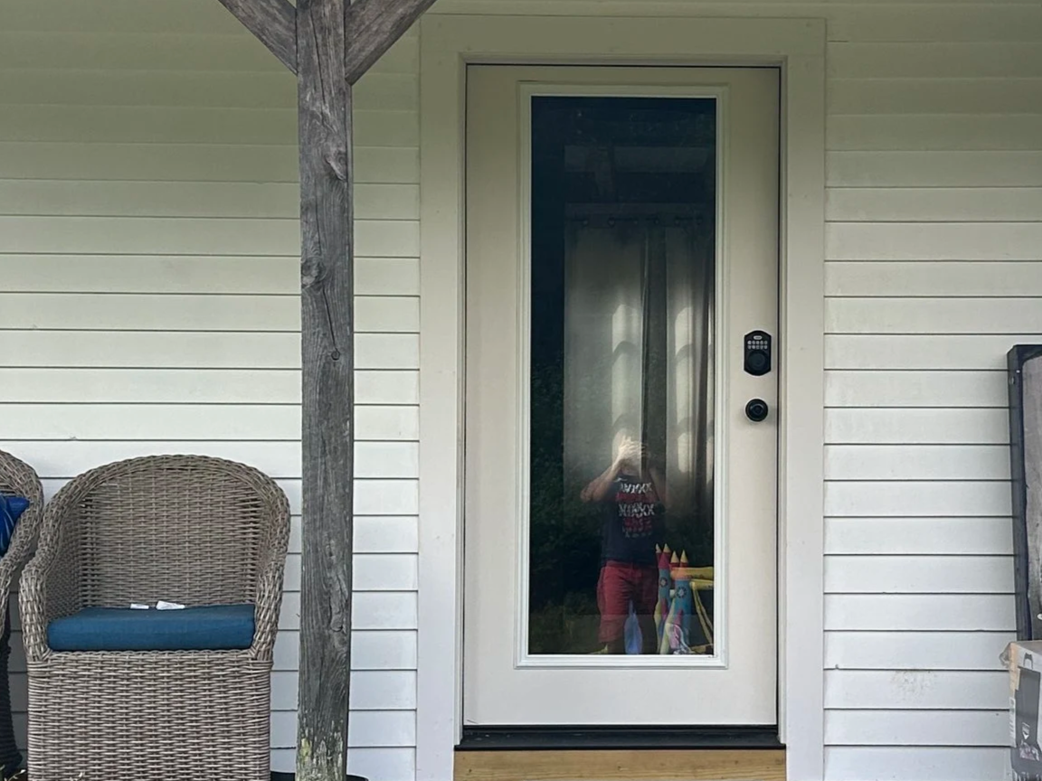 Residential house door with glass panel, digital keypad, and doorbell, flanked by wicker chairs and wooden support on a white siding exterior.