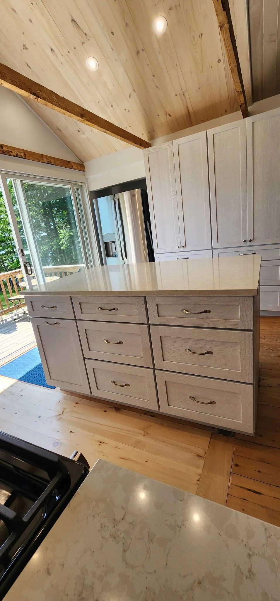 A modern kitchen with light wood cabinets, a large kitchen island with drawers, stainless steel refrigerator, open sliding glass door leading to an outdoor deck with furniture, and a wood-paneled vaulted ceiling with recessed lighting.