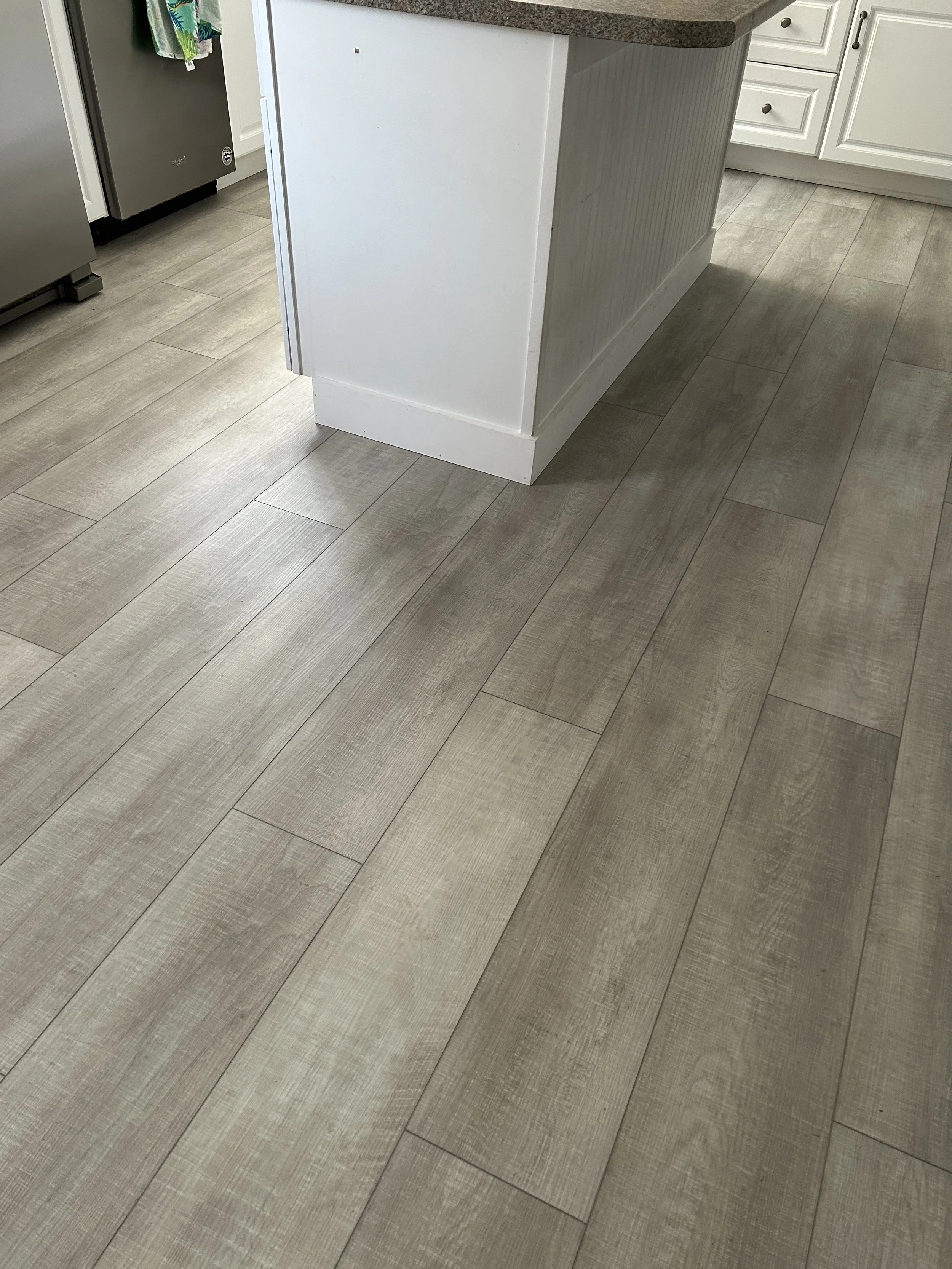 Photo of a kitchen floor with light gray vinyl flooring, a white kitchen island with a gray granite countertop, and white cabinets with black knobs. Part of a food refrigerator with colorful towels hanging on its door is visible on the left.