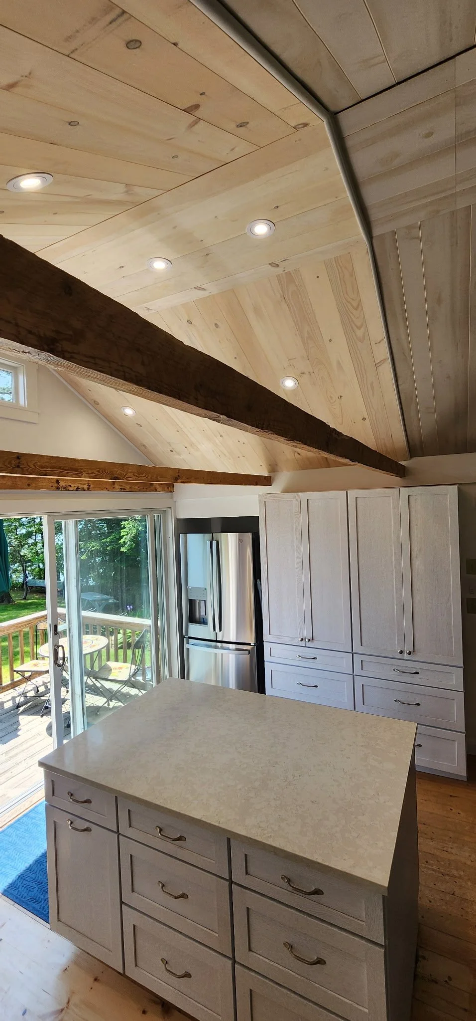 Kitchen with white cabinets, stainless steel refrigerator, island with beige countertop, wood beam ceiling, sliding glass door leading to outdoor patio with table and chairs, and a view of green trees outside.