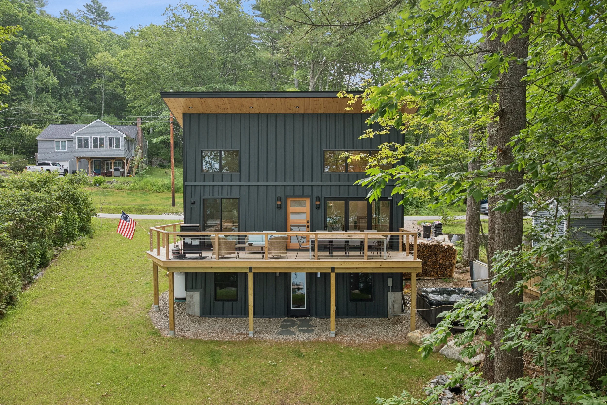 The image shows a modern, dark-colored house with a wooden deck in a lush green, wooded area. In the background, there are other houses and trees. The deck has outdoor furniture and an American flag.