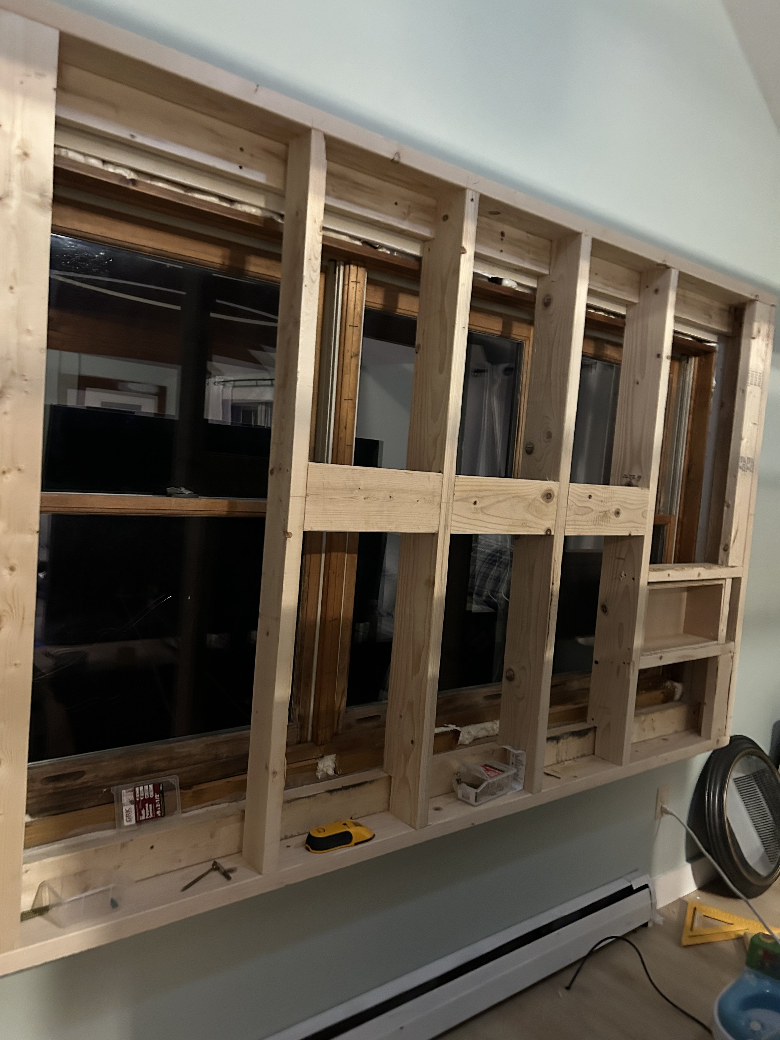 Partially constructed wall tv frame with exposed wooden studs and a window, tools, and construction materials in a room.