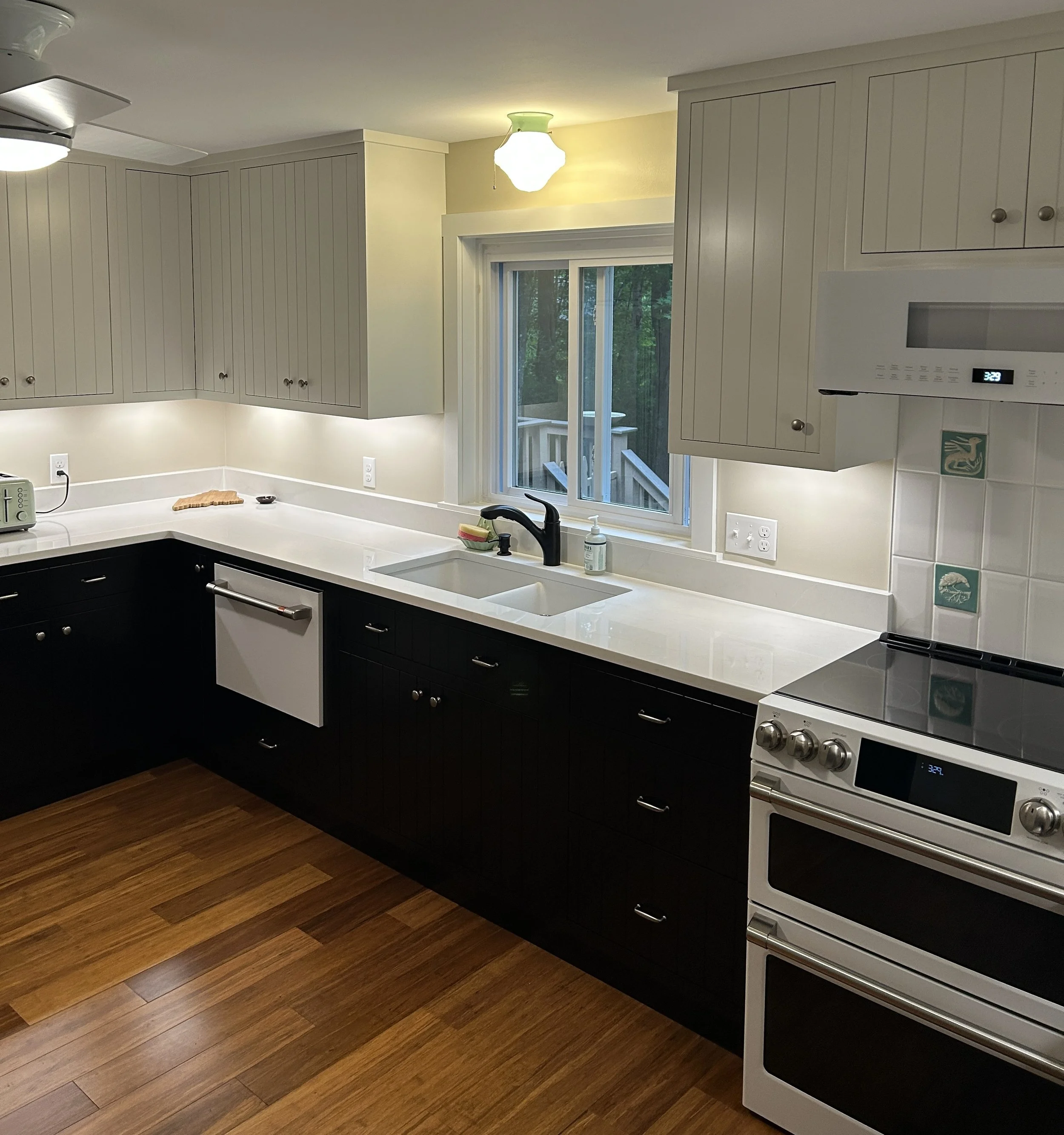Kitchen with white upper cabinets, dark lower cabinets, a white countertop, a stainless steel stove, a window with green foliage outside, and wood flooring.