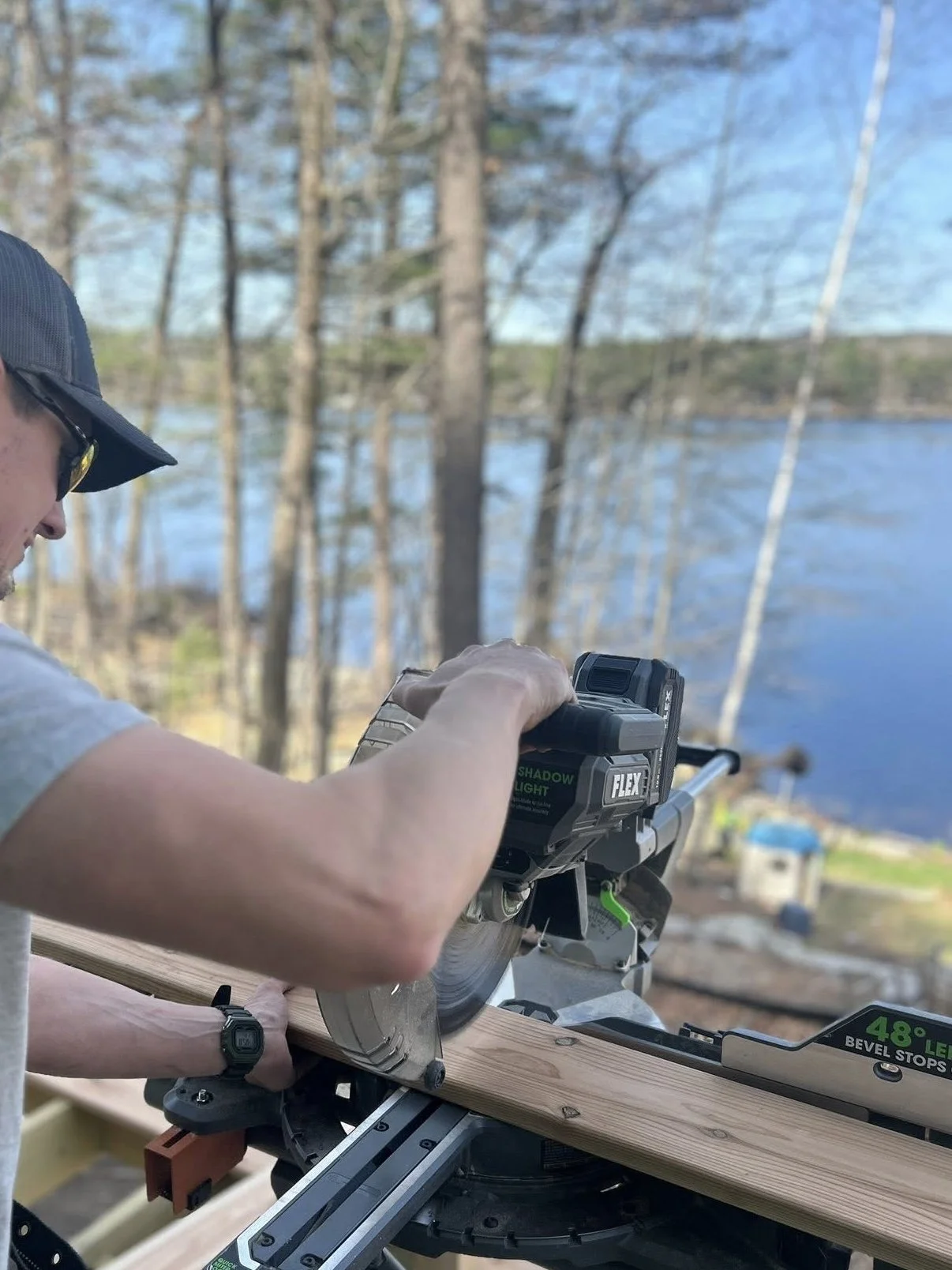 Avex Custom Homes owner Andrew is operating a power saw outdoors near a lake, with trees in the background. They are wearing a baseball cap, sunglasses, and a wristwatch, cutting a piece of wood on a chop saw. 